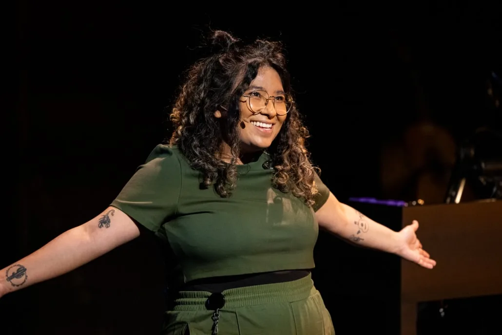 Satya Chavez, wearing a green body suit, smiling and arms wide open on stage.