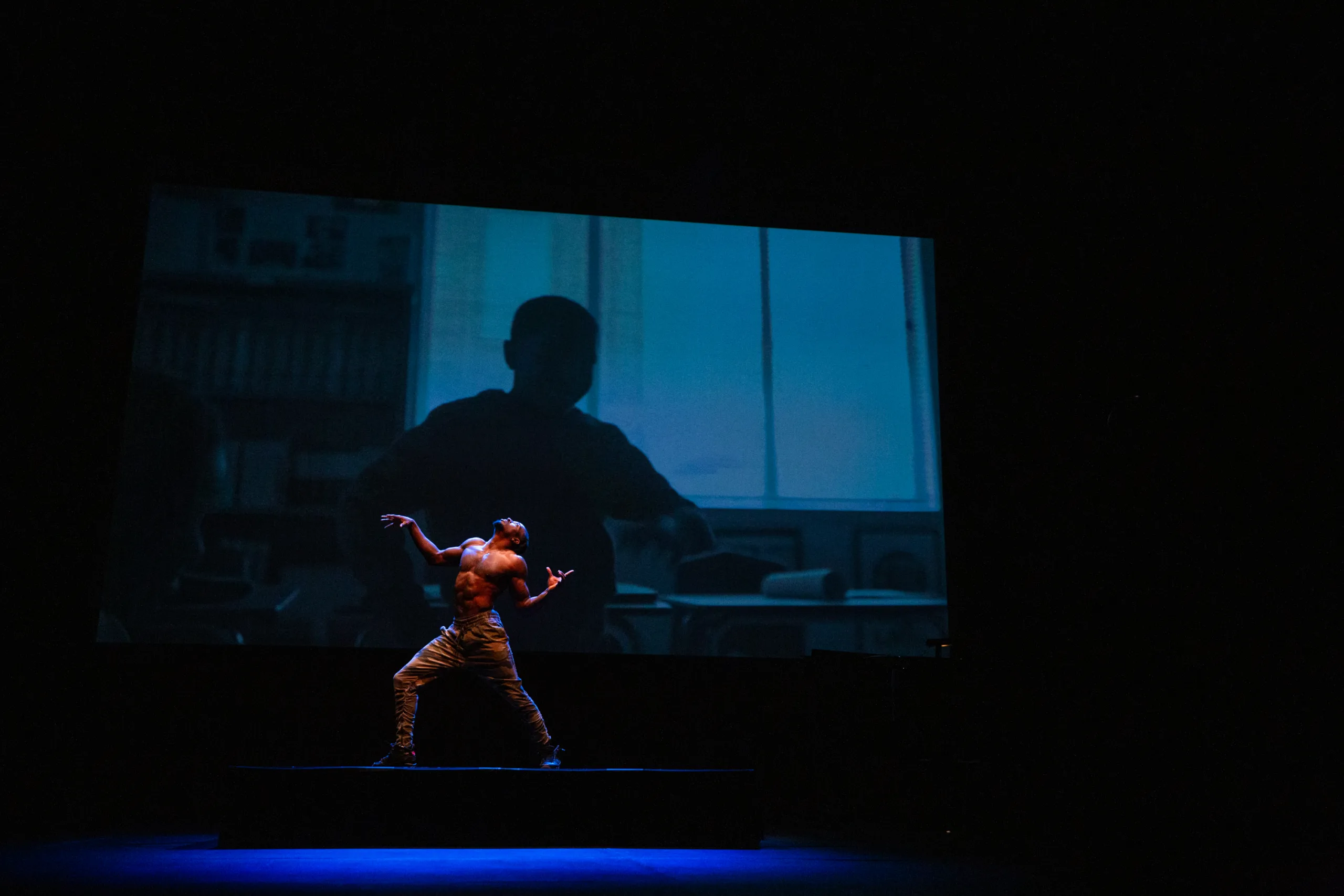 Man dancing on stage shirtless in front of a projection of a young boy in the background.