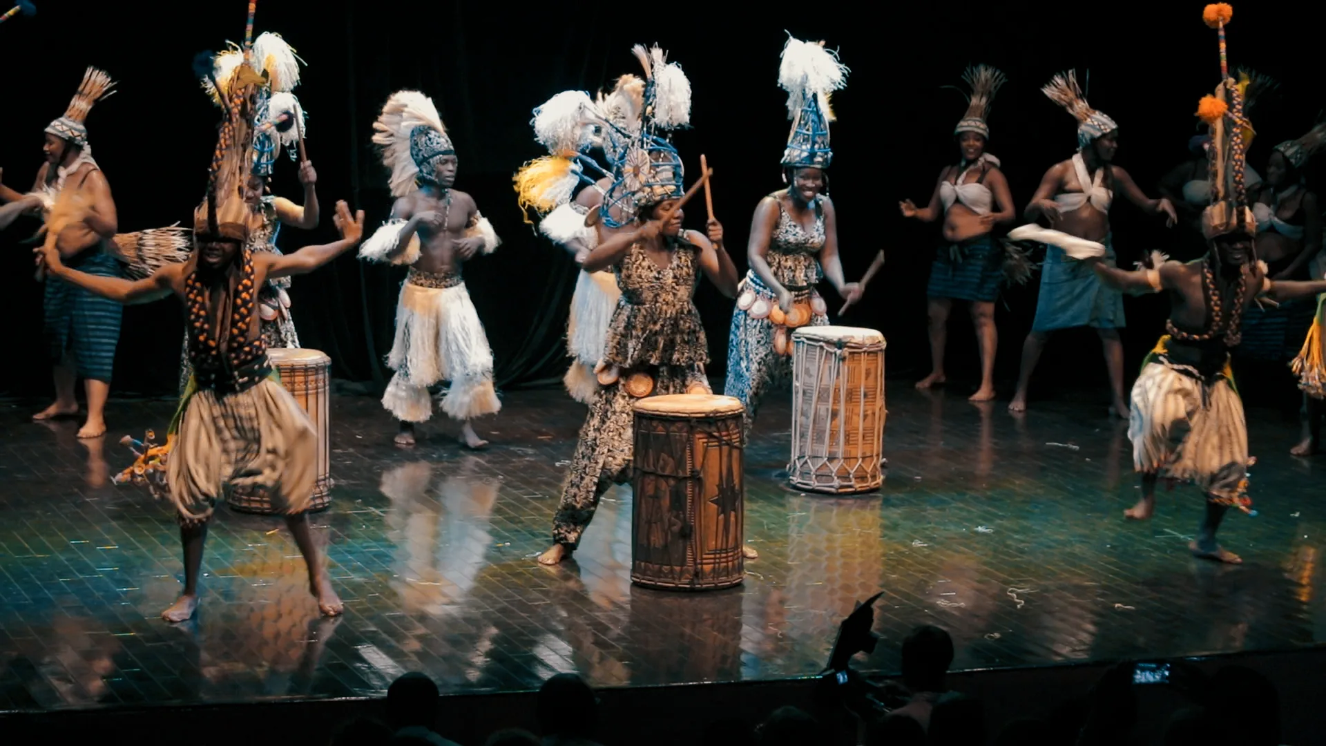 The company of Les Ballets Africains on stage in costume dancing in formation.