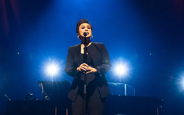 Lea Salonga performs on stag singing with microphone and stage lights behind her.