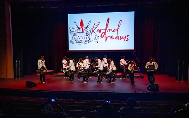 Video screen on stage reading Kardinal dreams and students performing with drums.