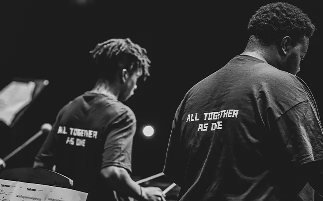 close up shot of students from behind drumming with t-shirts that read all together as one.