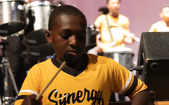 close up shot of student wearing yellow synergy shirt and drumming.