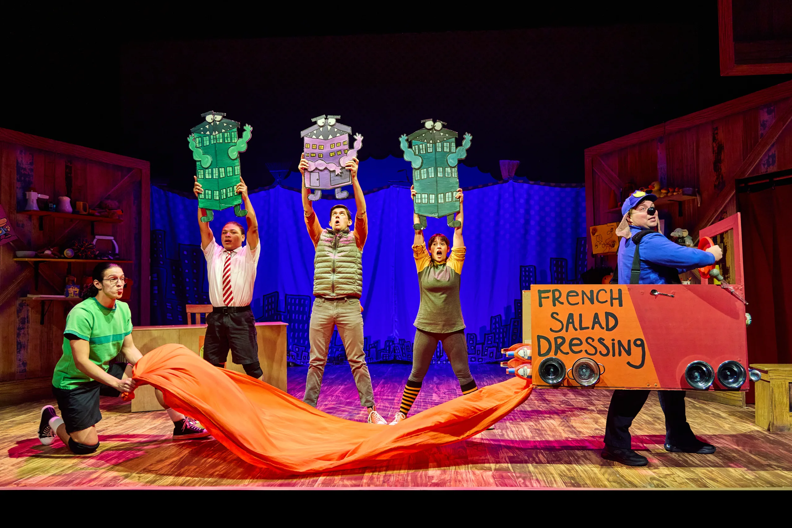 The cast of Dog Man on stage with five actors holding signs with Dog Man driving a cardboard truck with 