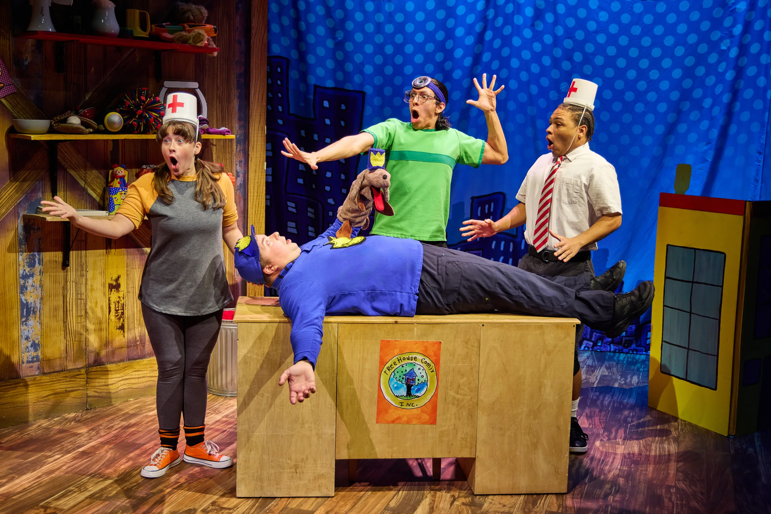 The cast of Dog Man on stage with three actors wearing Doctor and Nurse accessories and the postman lying on the table with a puppet.