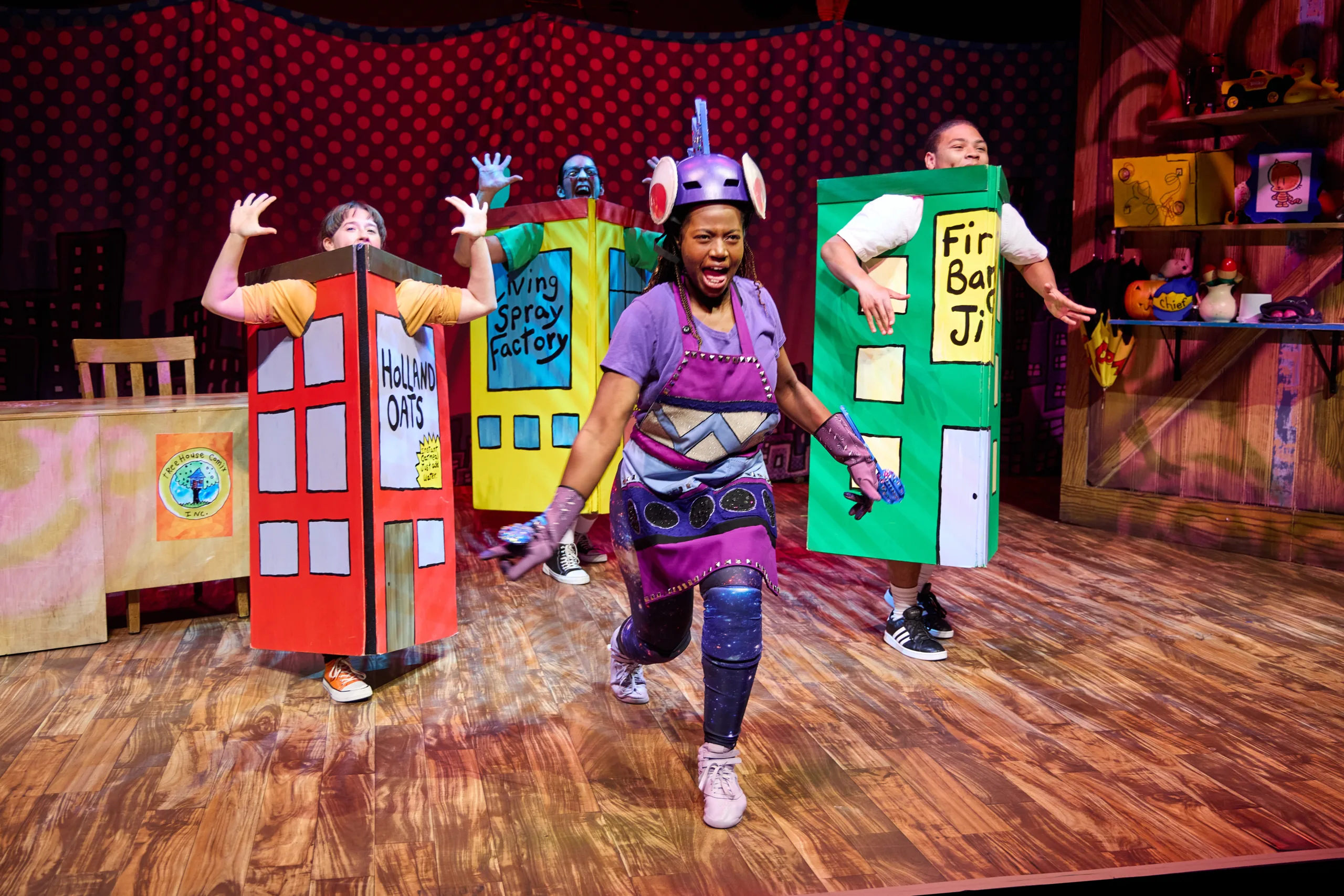 The cast of Dog Man on stage with three actors in cardboard costumes of buildings with a purple costumed character running in the middle.