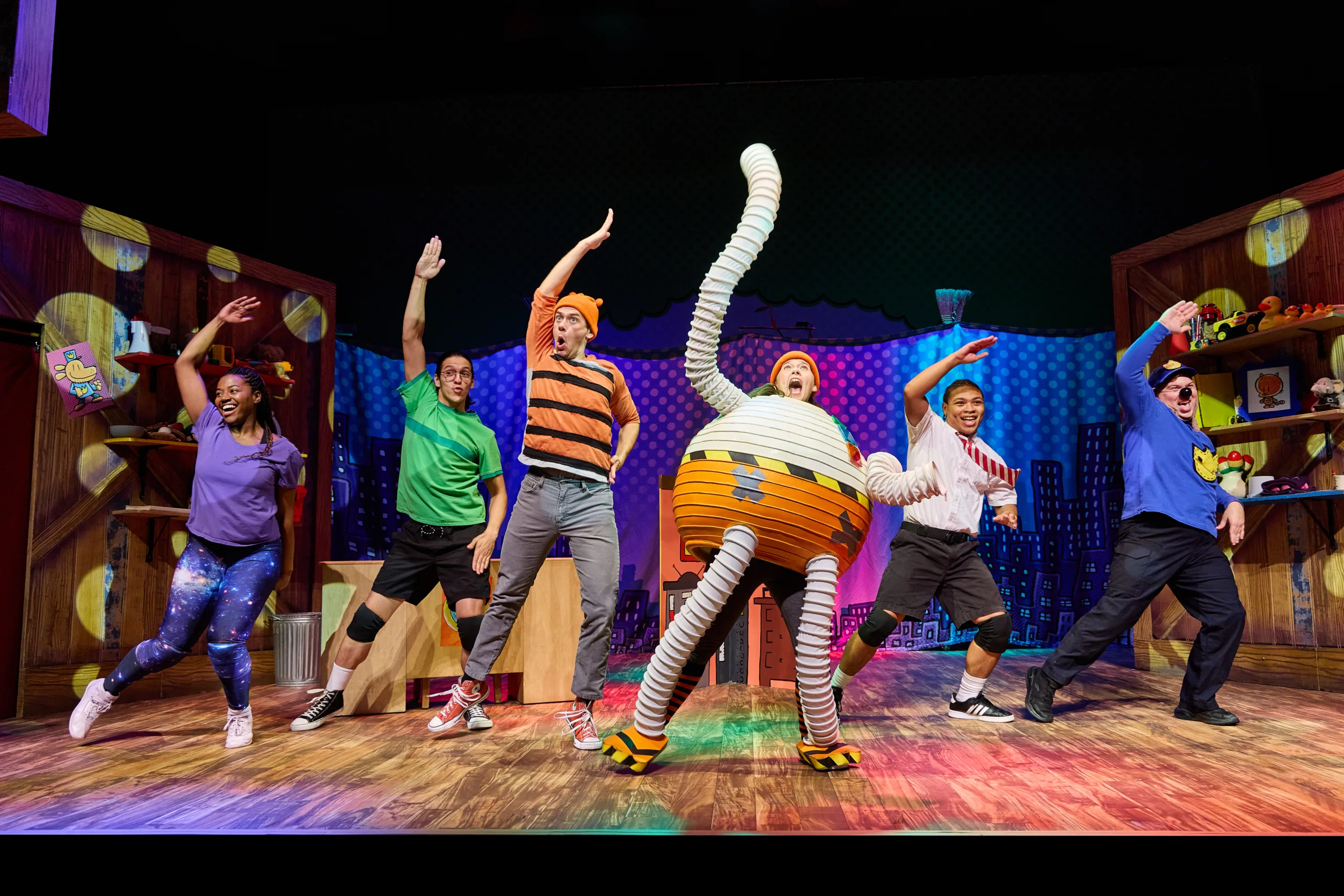 The cast of Dog Man on stage with six actors in costume pointing stretching their right arm over their heads.