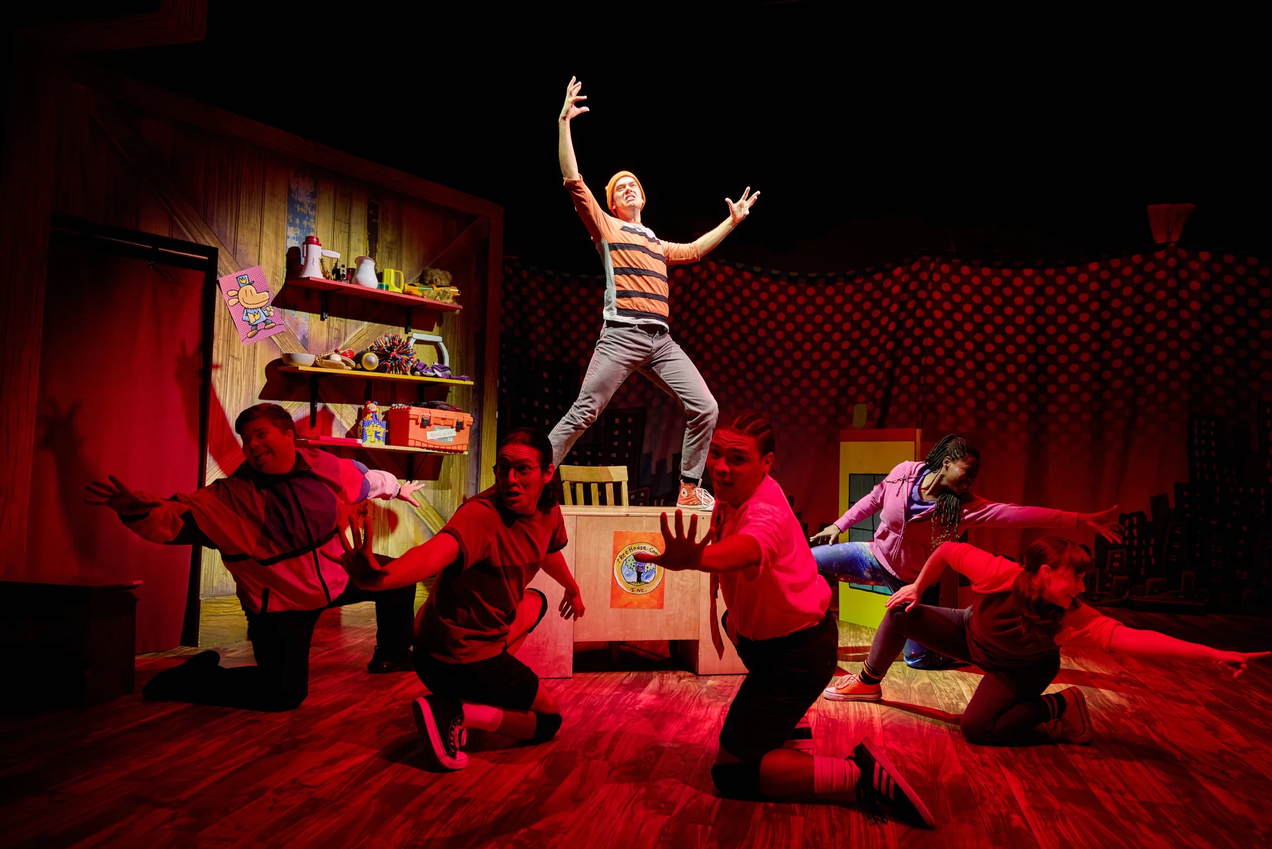 The cast of Dog Man on stage with five actors in kneeling in red light with arms outstretched and an actor standing on a table in the middle wearing a striped orange shirt.
