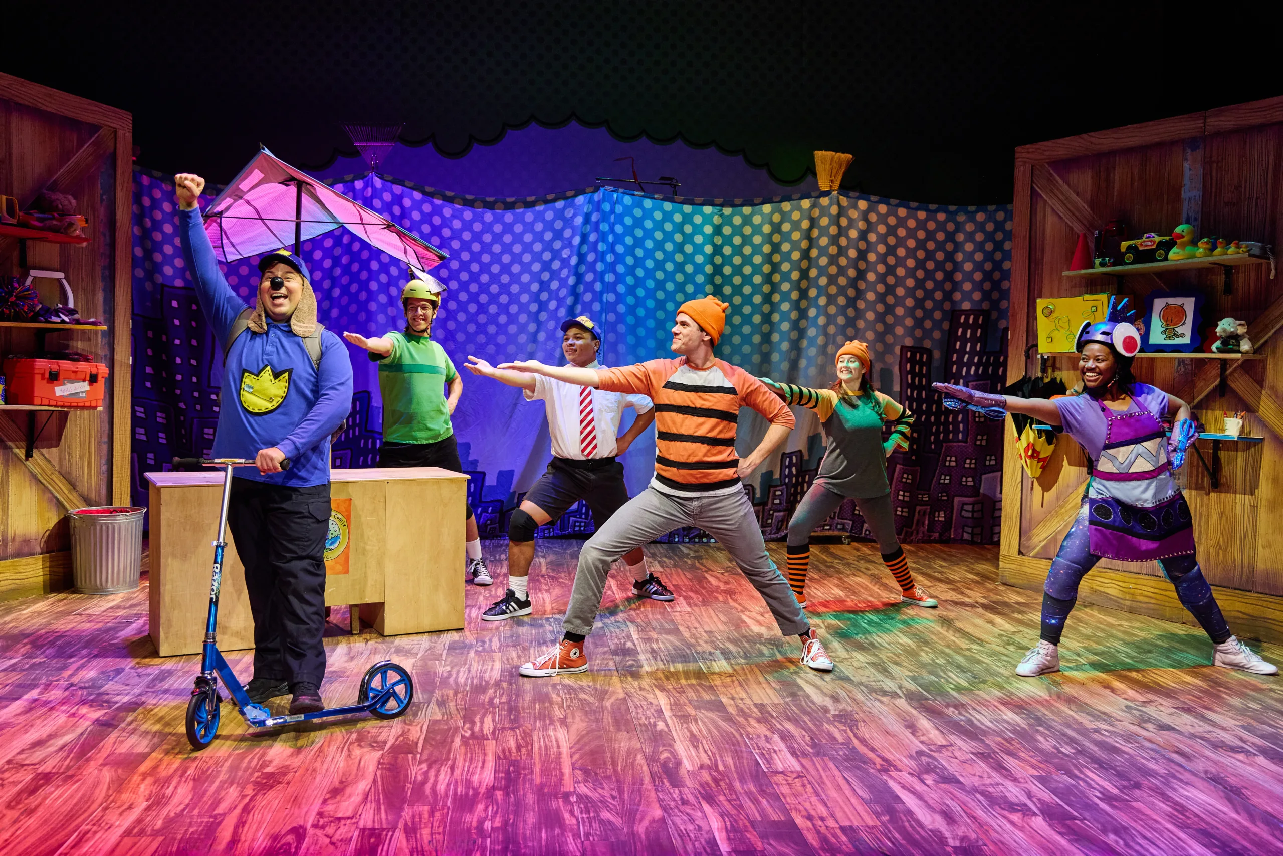 The cast of Dog Man on stage with five actors in costume pointing toward Dog Man on a scooter.