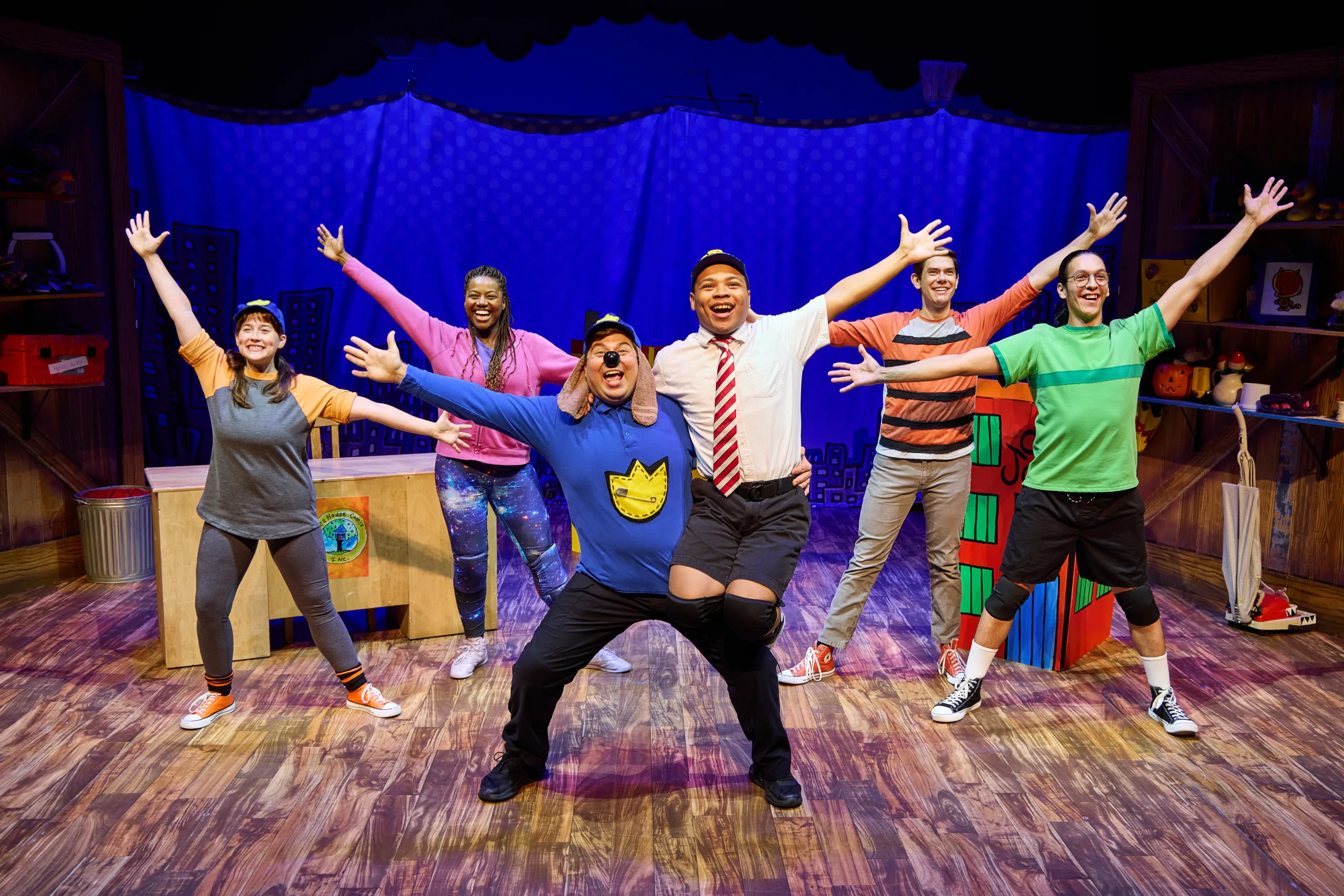 The cast of Dog Man on stage with six actors in costume posing with arms out with Dog Man in the middle balancing another actor on his knee.