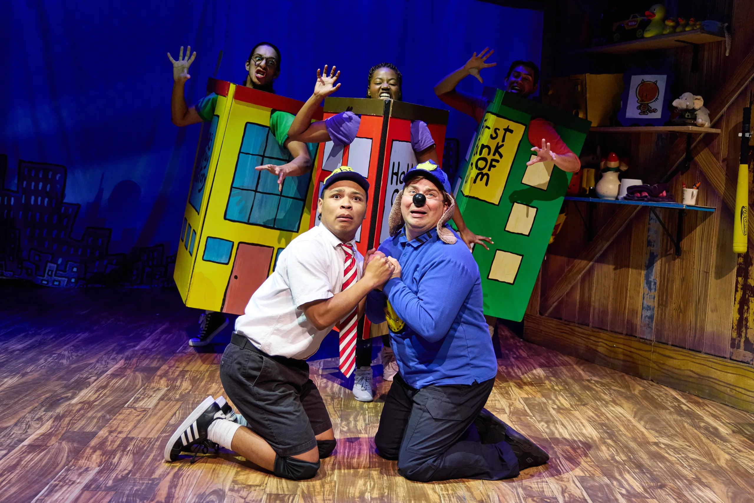 The cast of Dog Man on stage with three actors in cardboard costumes of buildings with Dog Man and another actor kneeling in the middle.