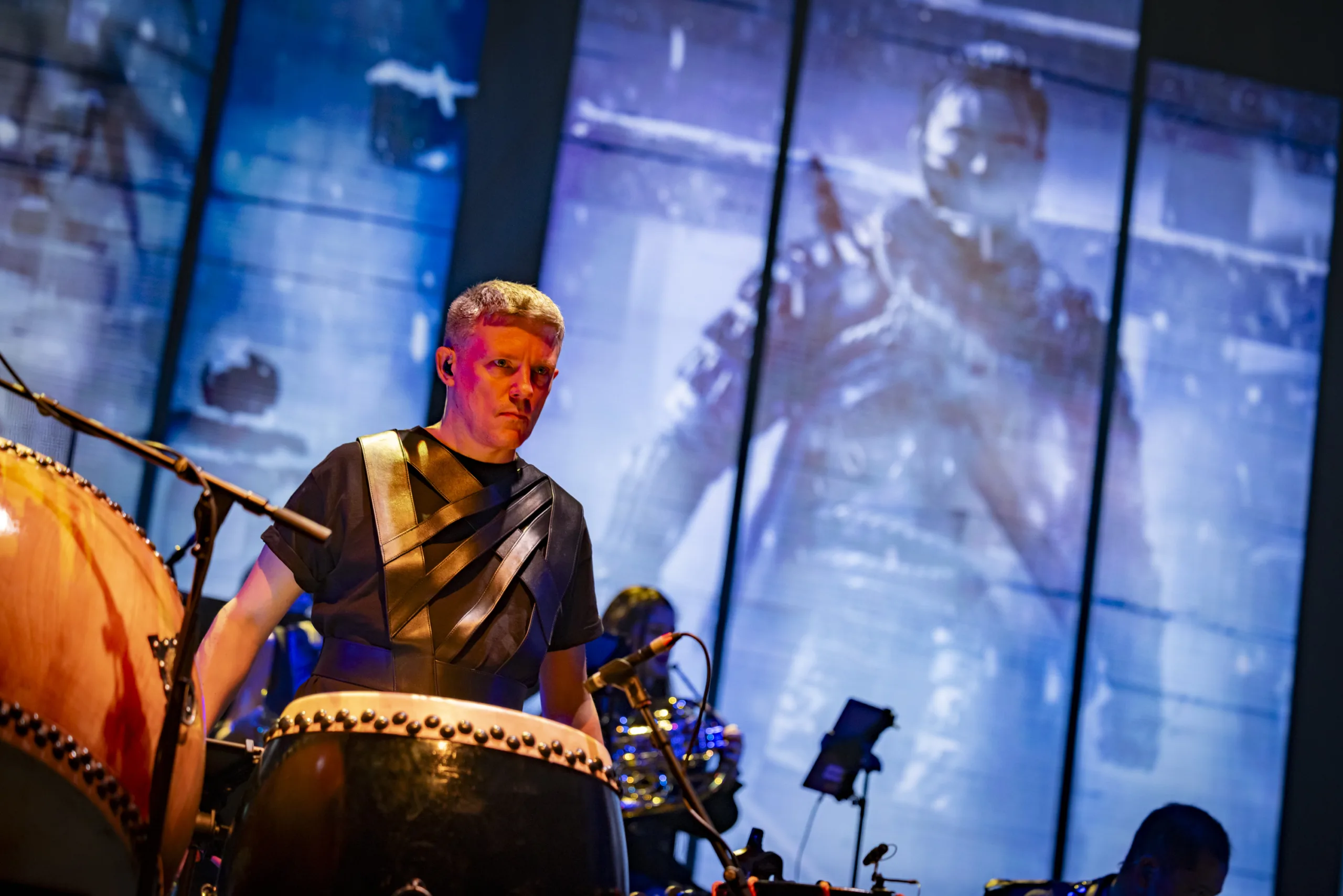 Close up of a drummer on stage in soft orange light with projections from 