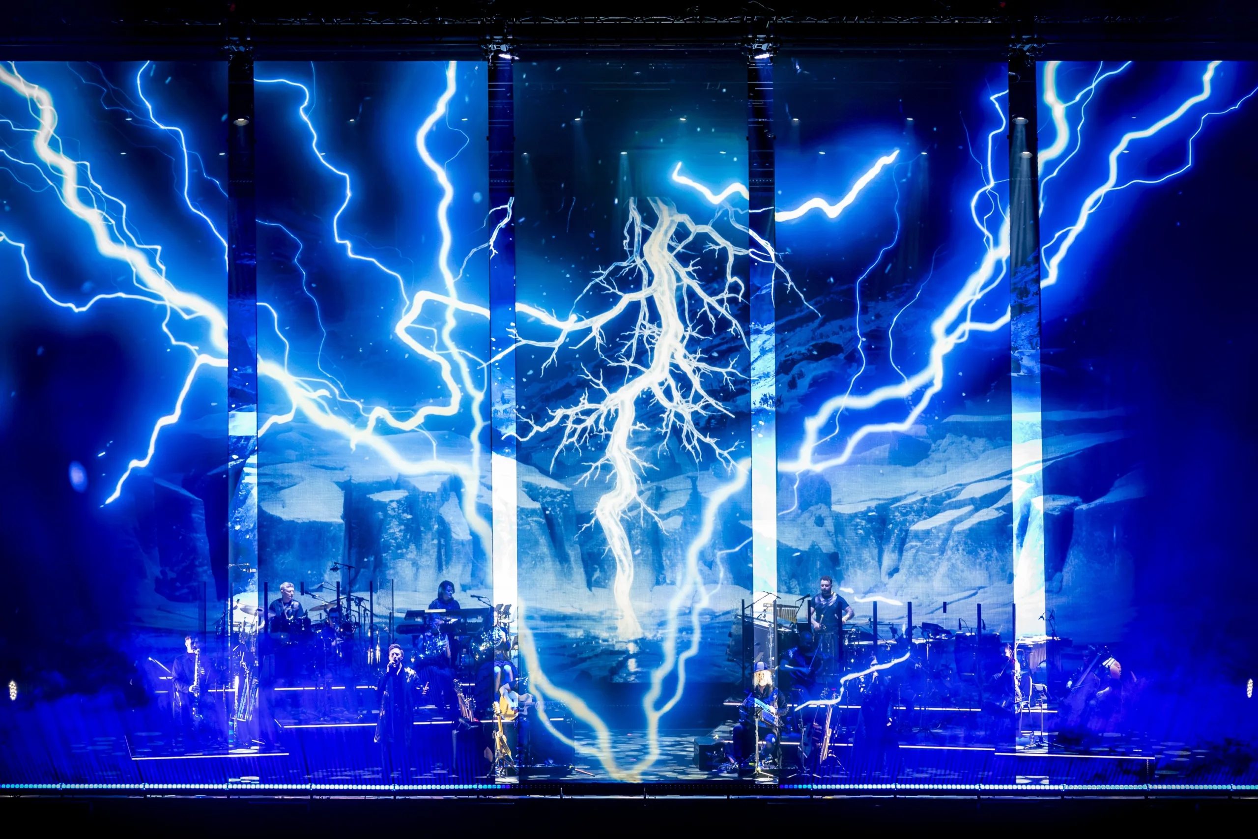 The band on stage in front of a projection of blue lightning strikes from 