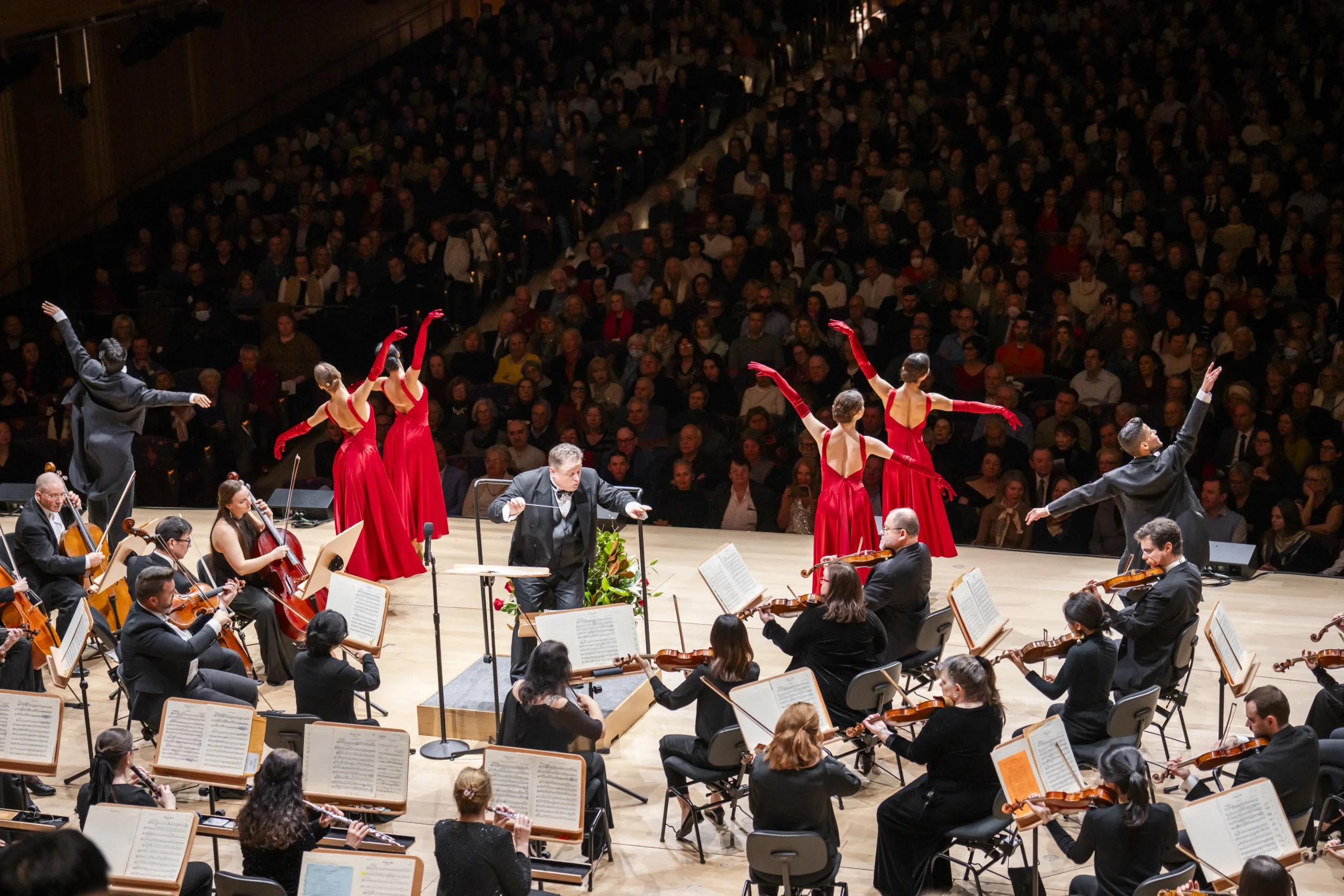 Salute To Vienna ballroom dancers from behind in red gowns posing with the orchestra around them on stage.