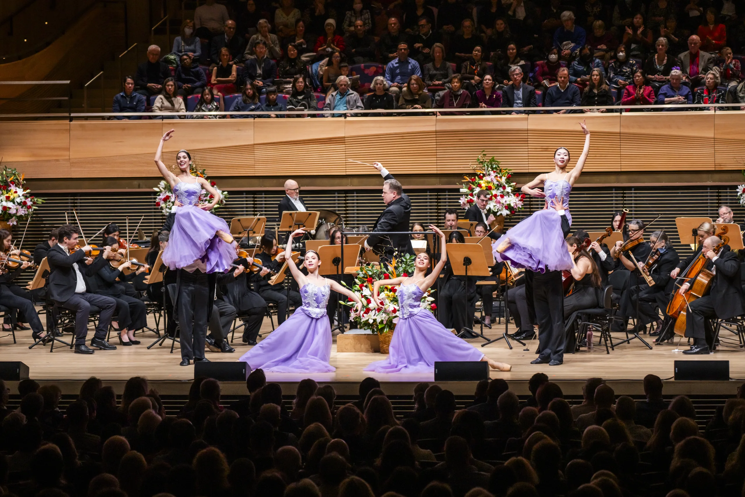 Salute To Vienna ballroom dancers in purple gowns posing with the orchestra around them on stage.