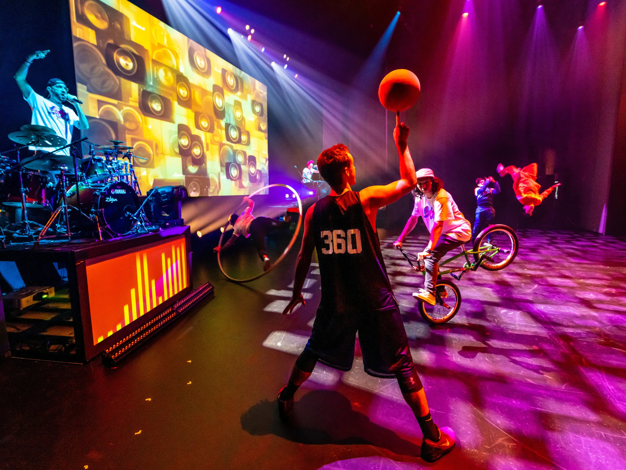 360 Allstars holding basketball, someone riding a BMX bike, beatboxing, acrobatics and drumming on stage.