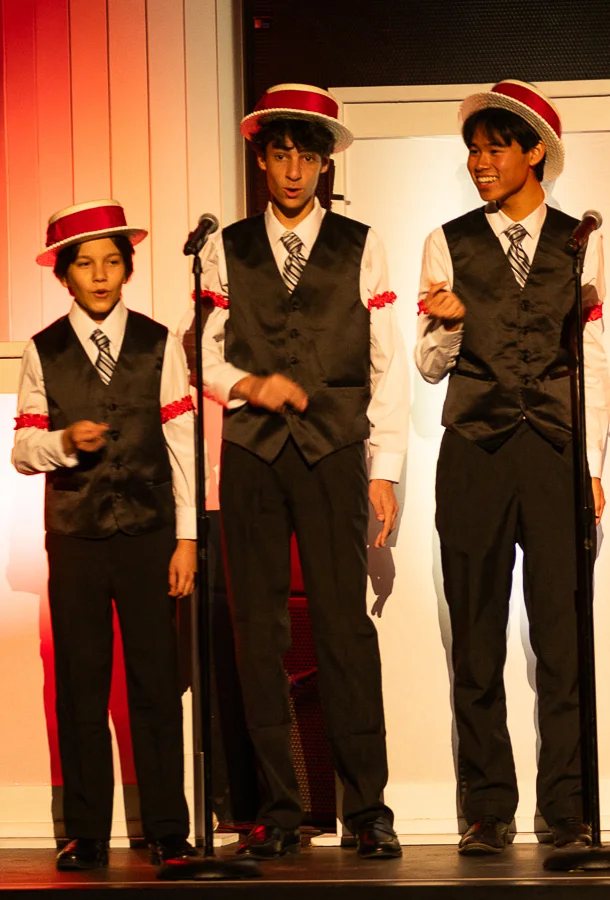 Three boys wearing barber shop-type outfit singing into one microphone.