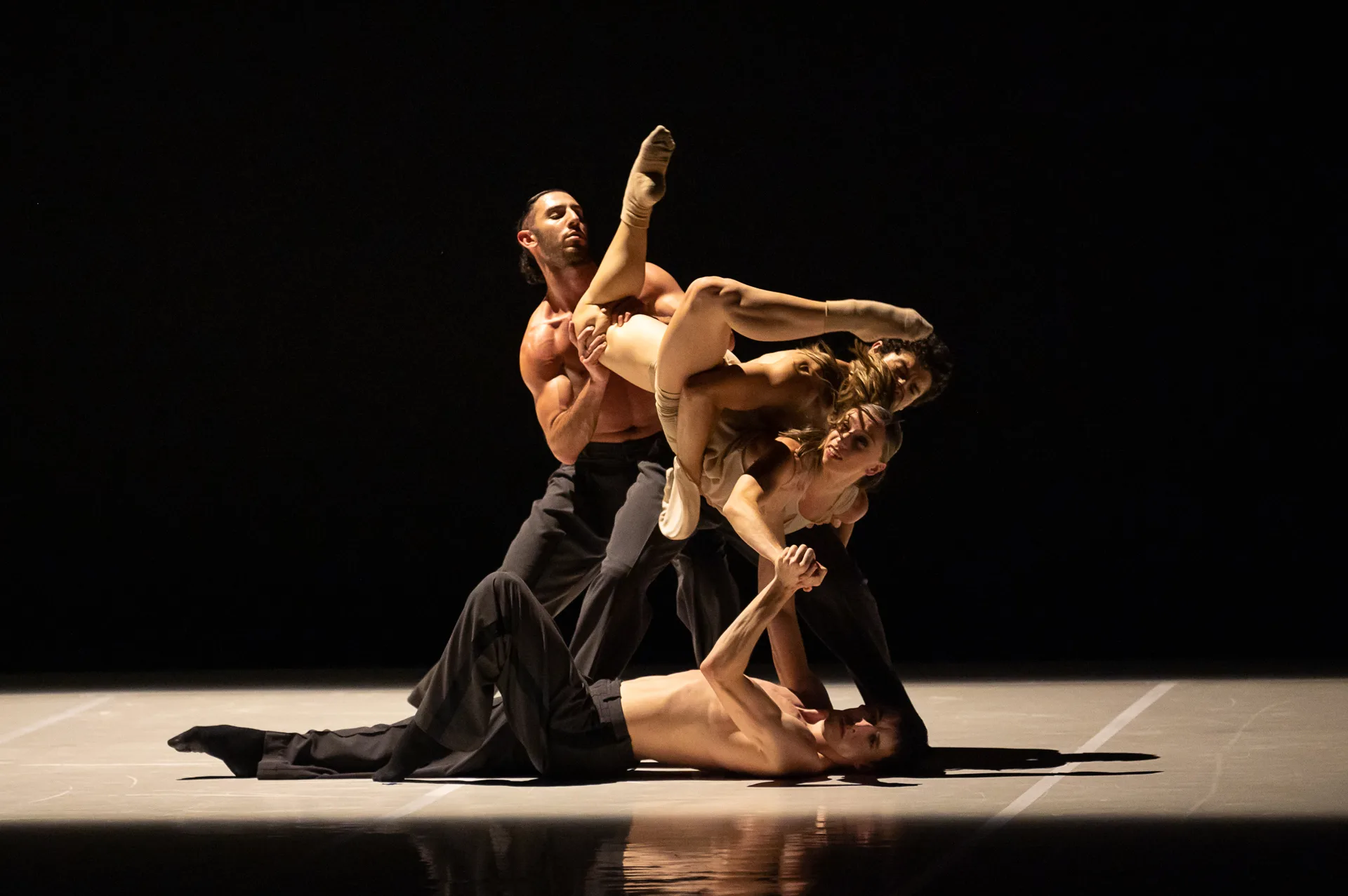 4 members of the ballet company in black in a complicated contortion on stage.