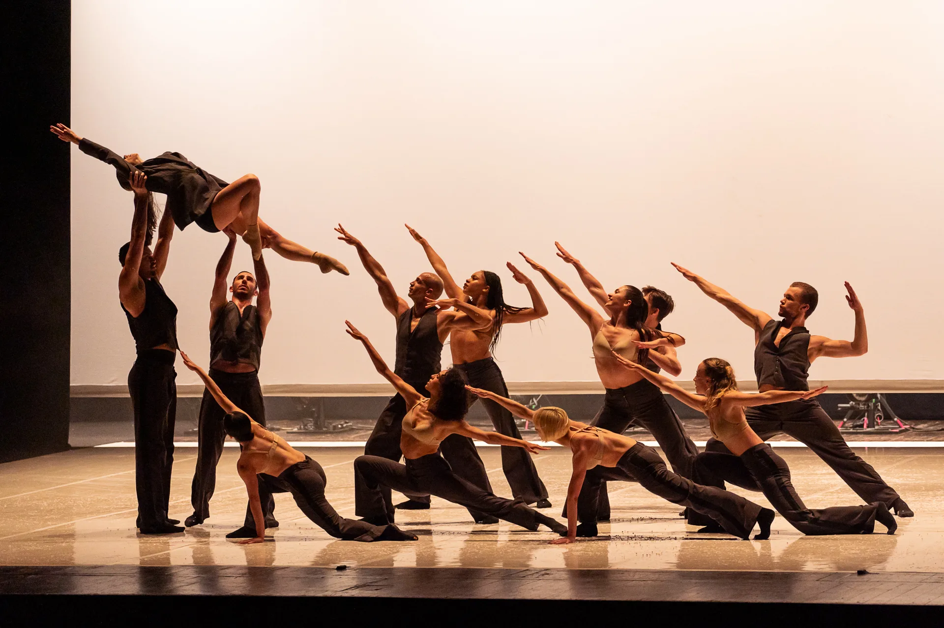 The ballet company in black and gold posing crouching toward one dancer being lifted to the left with their arms extended in soft light.