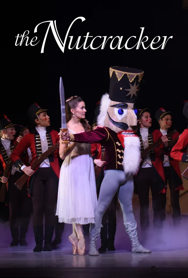 Scene from 'The Nutcracker' featuring a Nutcracker character with a sword beside a ballerina in white, with performers in red and black uniforms in the background.