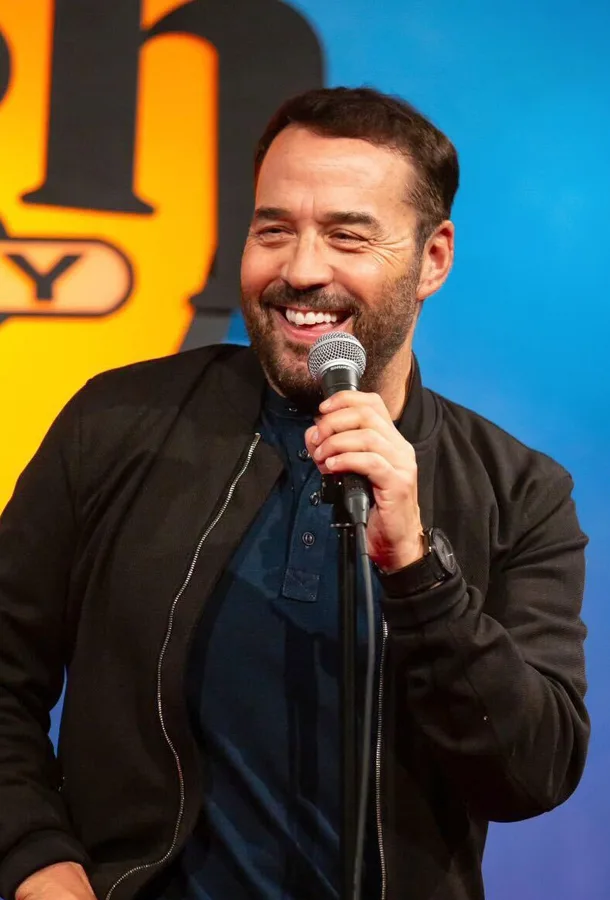 Jeremy Piven holds up microphone and is wearing a blue shirt with a black zip up.
