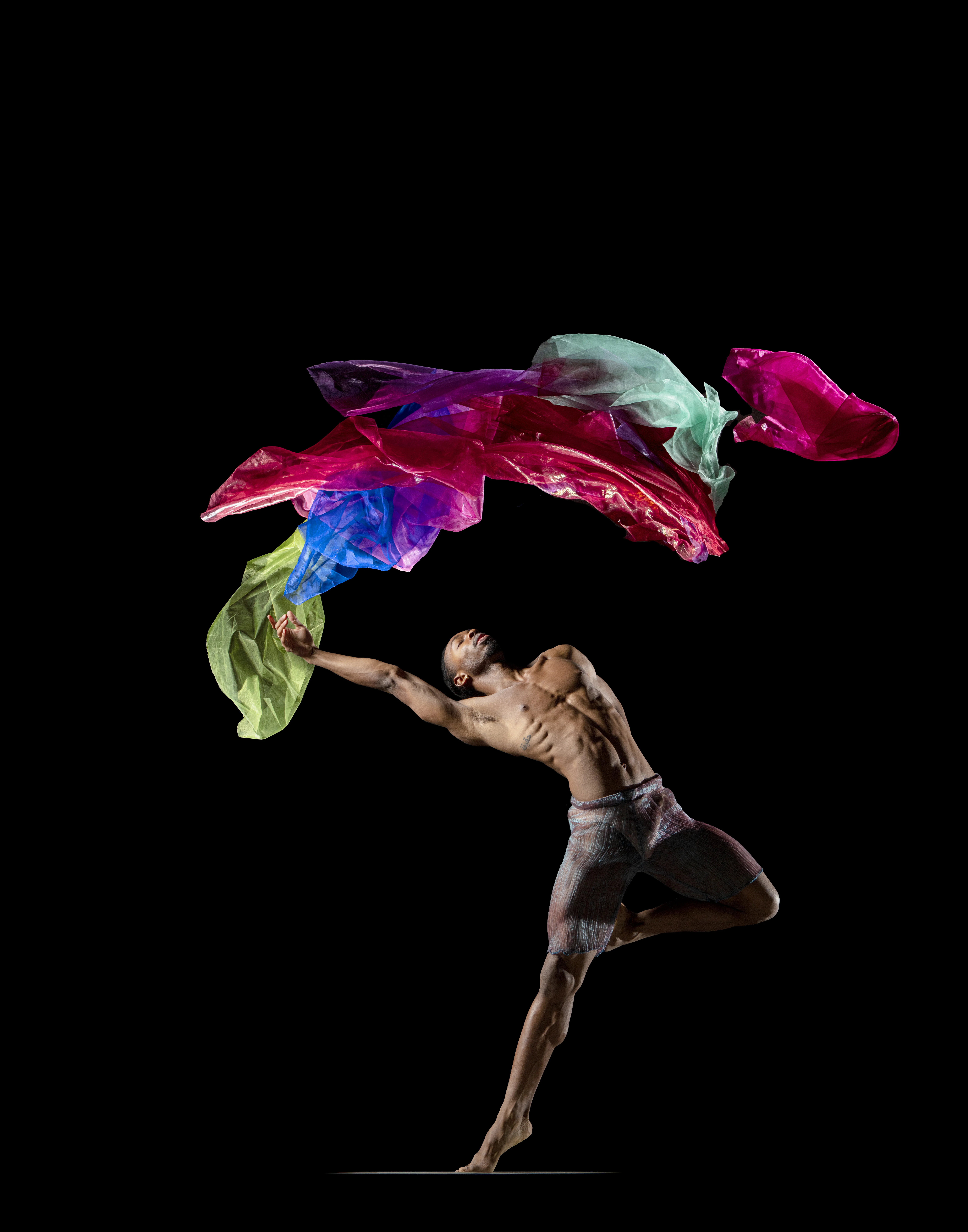 Dancer in dynamic pose reaching toward colorful floating fabrics against a black background, wearing shorts and no shirt.