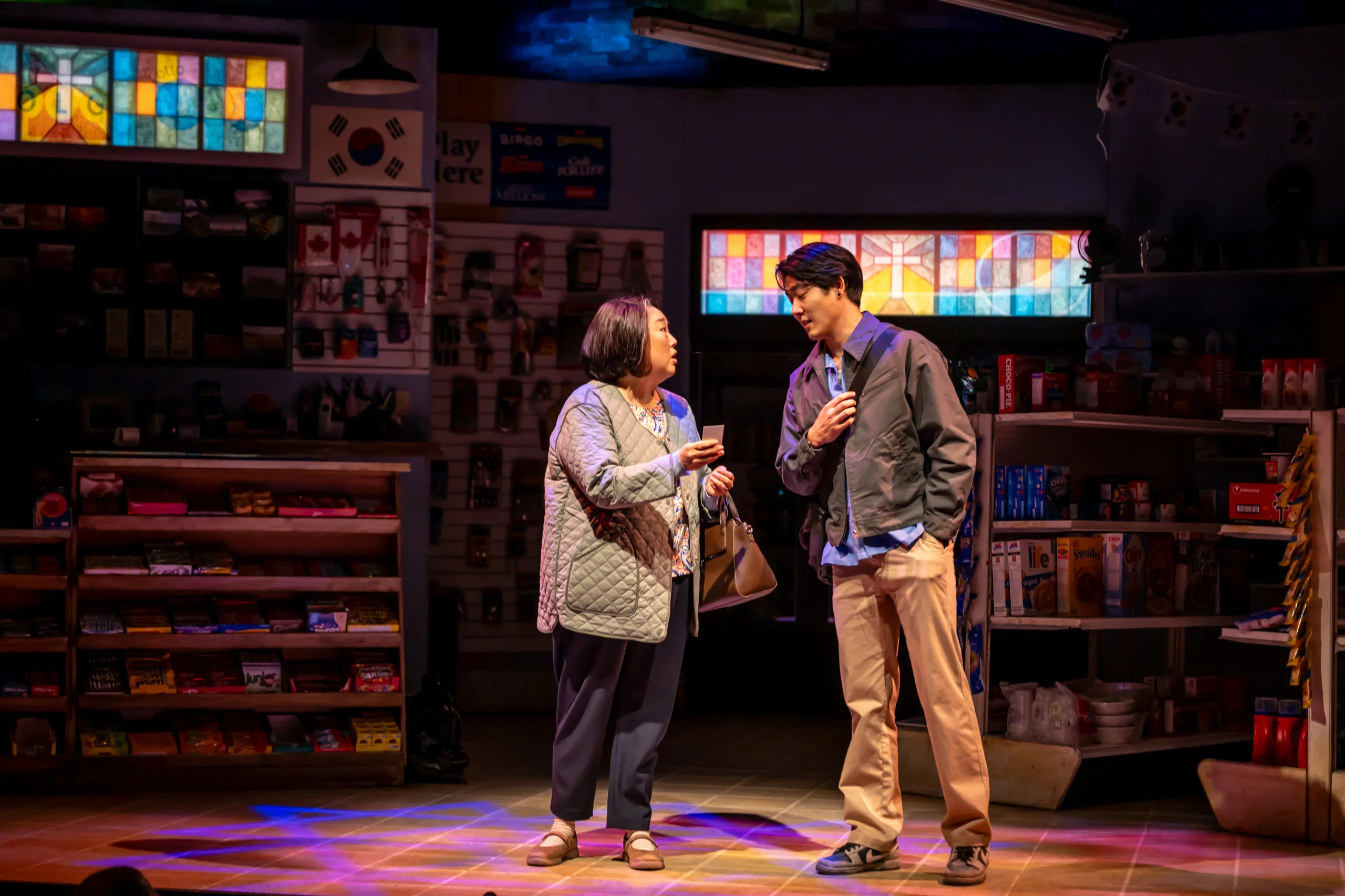 Two people standing in a darkly lit convenience store set. 