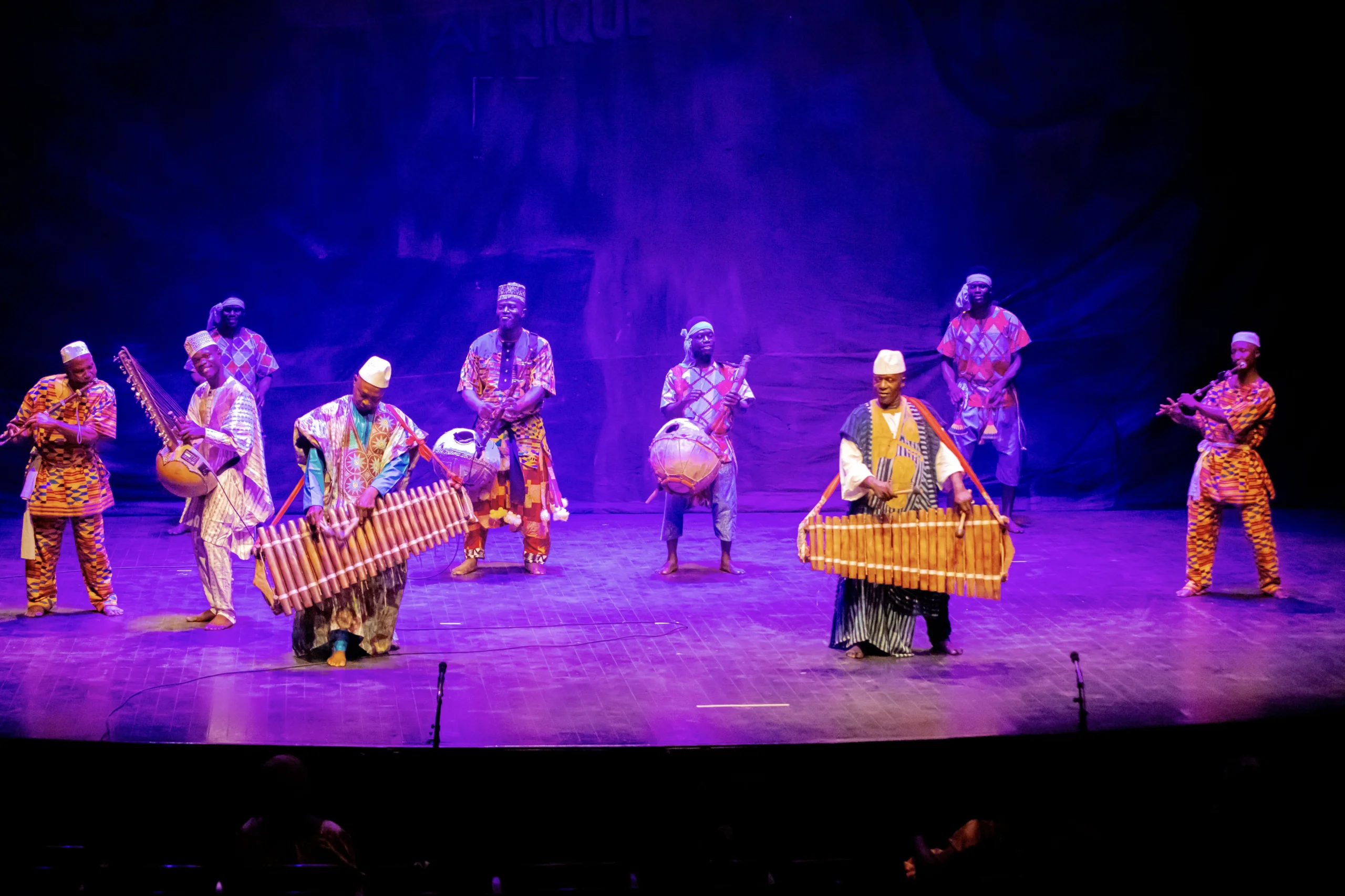 The company of Les Ballets Africains on stage in costume dancing in formation with instruments.
