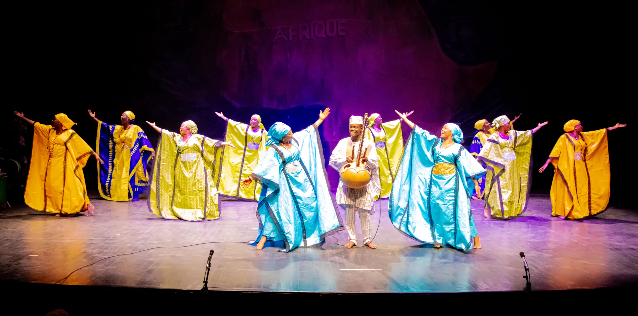 The company of Les Ballets Africains on stage in costume dancing in formation.