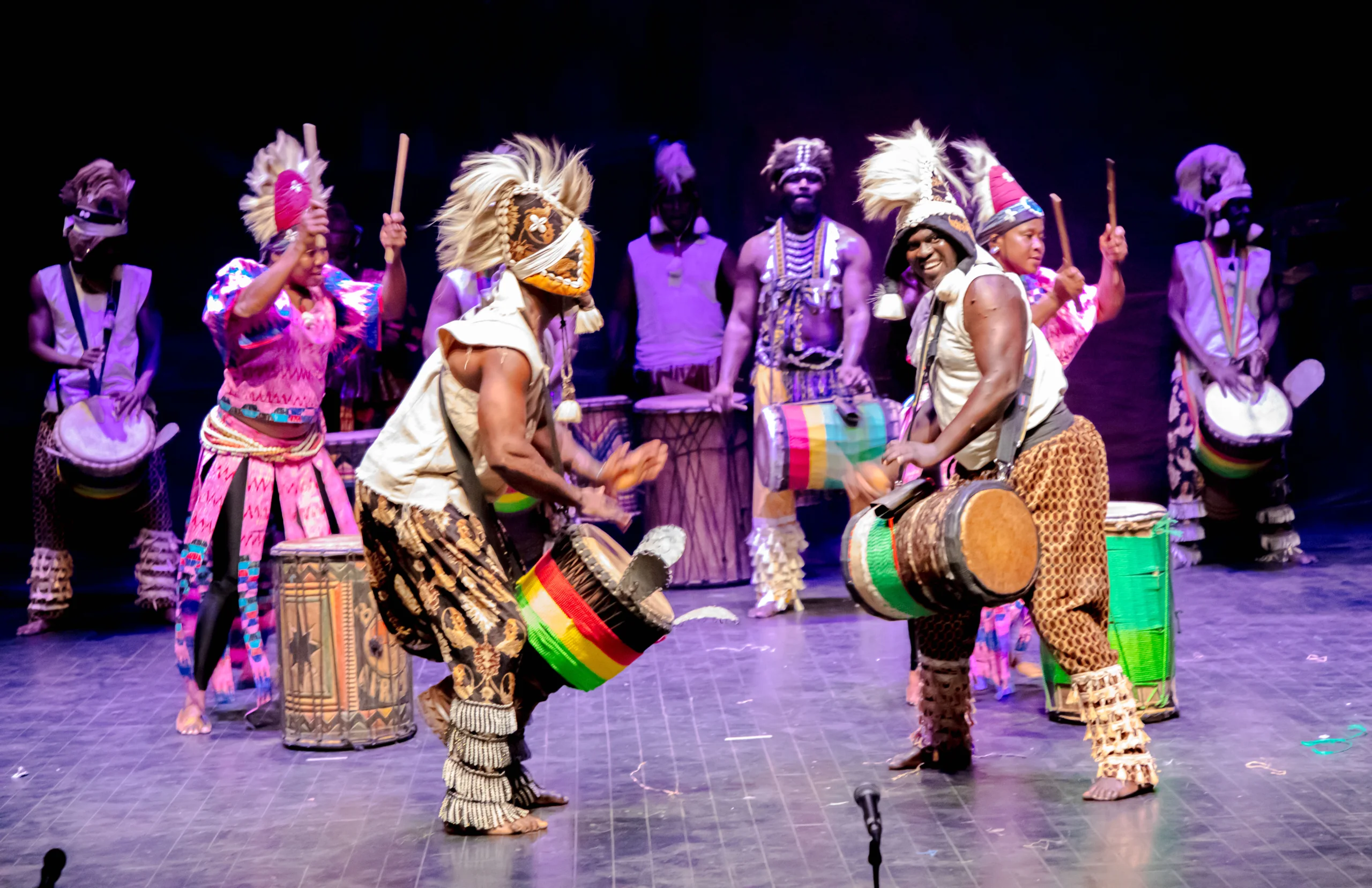The company of Les Ballets Africains on stage in costume dancing with drums.