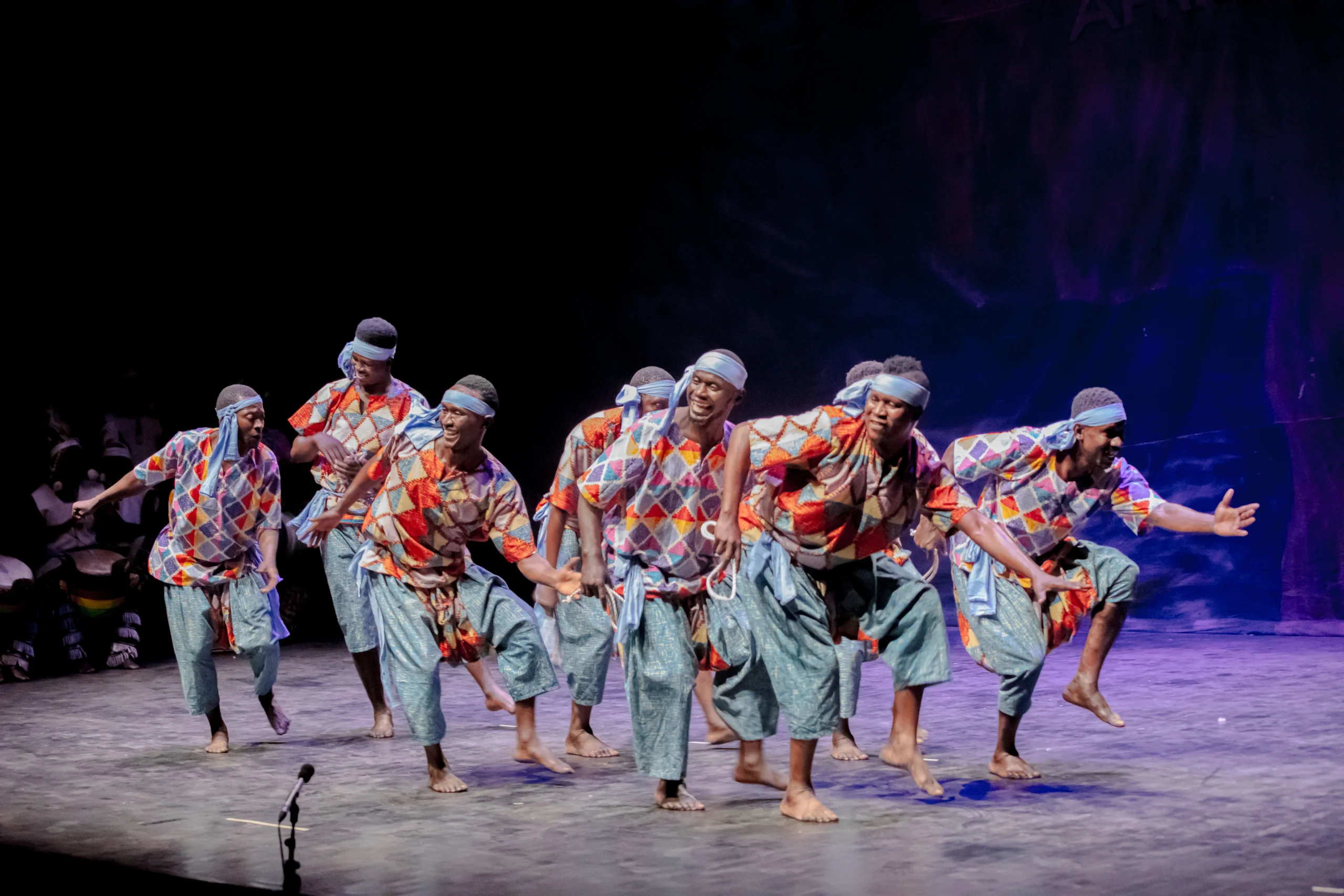 The company of Les Ballets Africains on stage in costume dancing in formation.