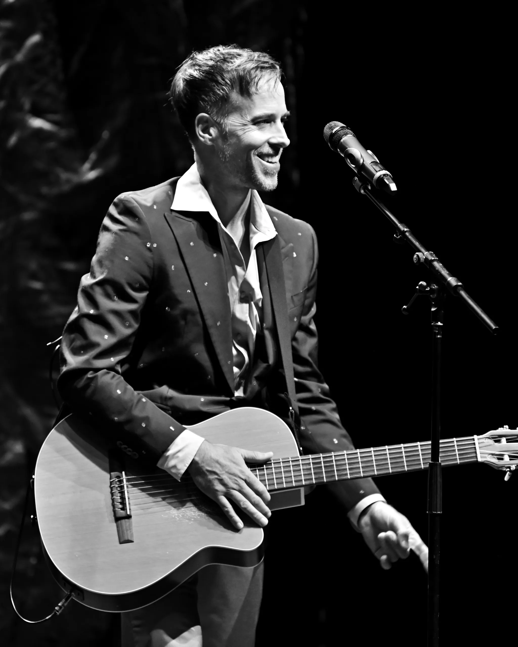 Luke McMaster smiling, holding a guitar at a microphone stand.