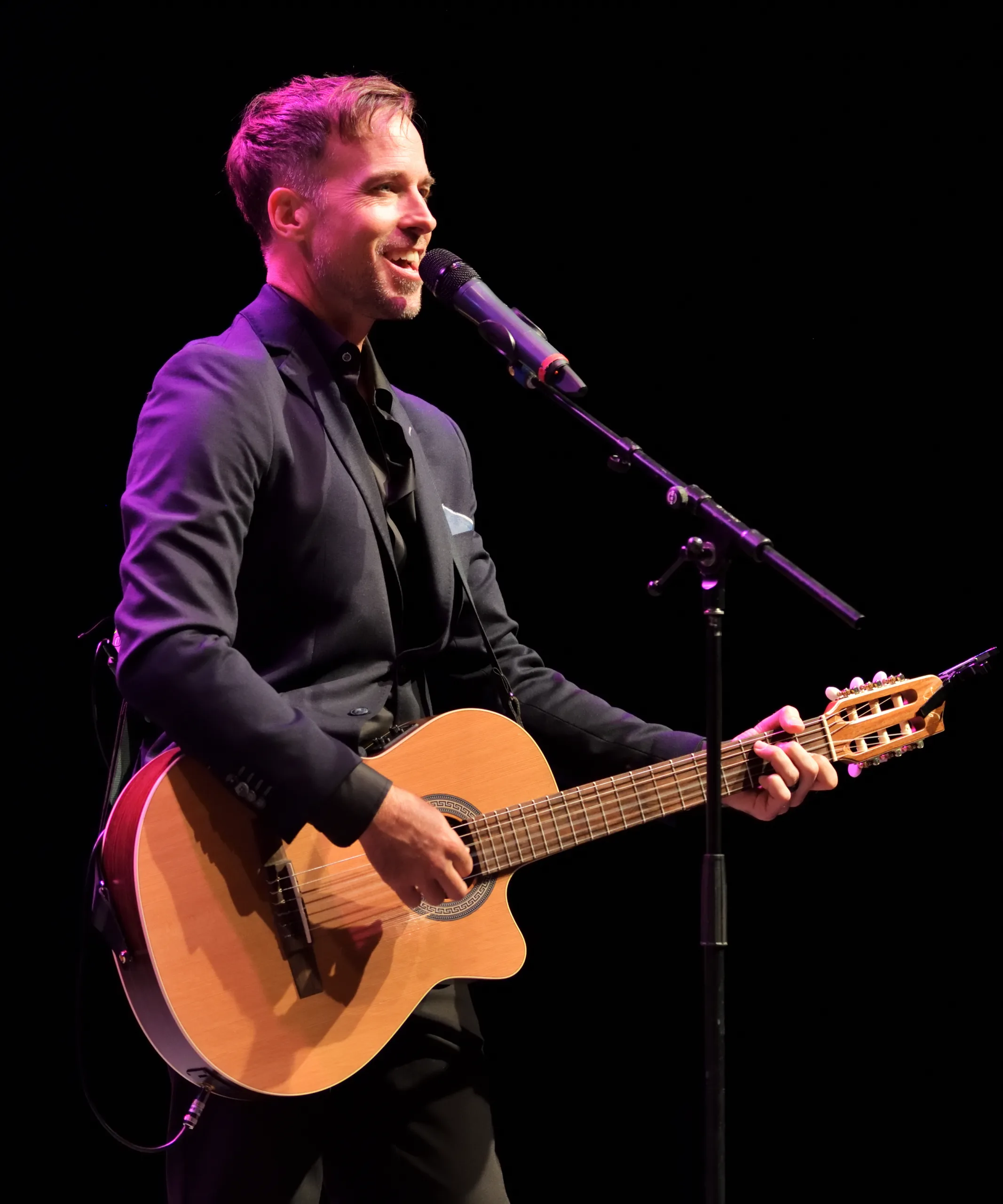 Luke McMaster smiling, holding a guitar at a microphone stand.
