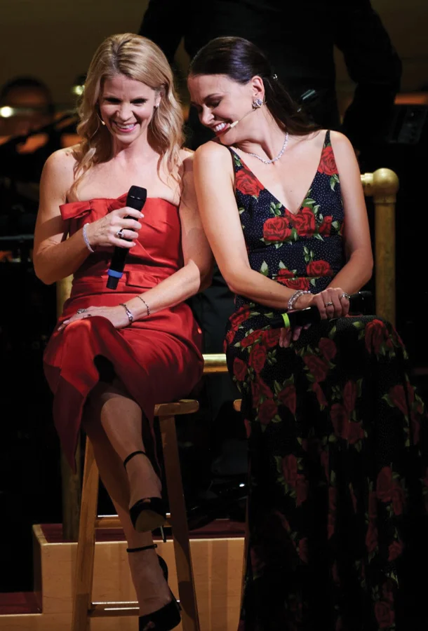 Kelli O'Hara and Sutton Foster holding microphones on stage in dresses leaning toward one another smiling.