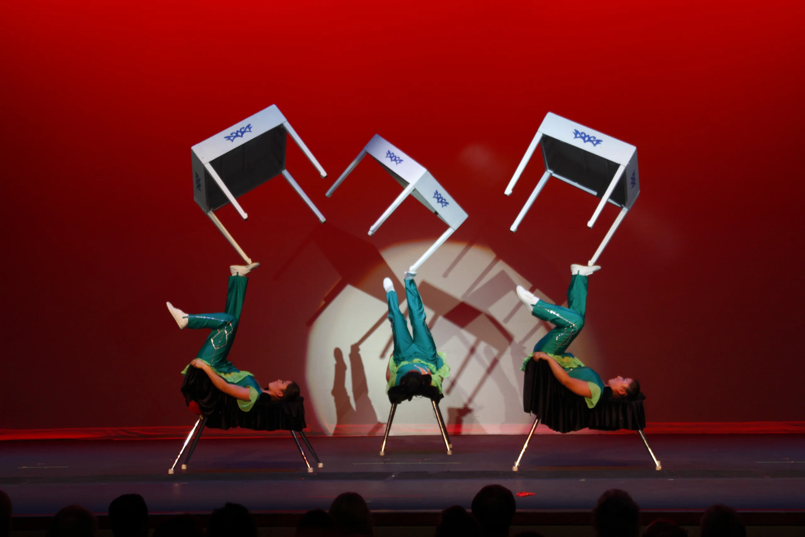 Three performers in green outfits lie on their backs on chairs on a red-lit stage, each balancing a tilted table in mid-air with their feet under a spotlight.