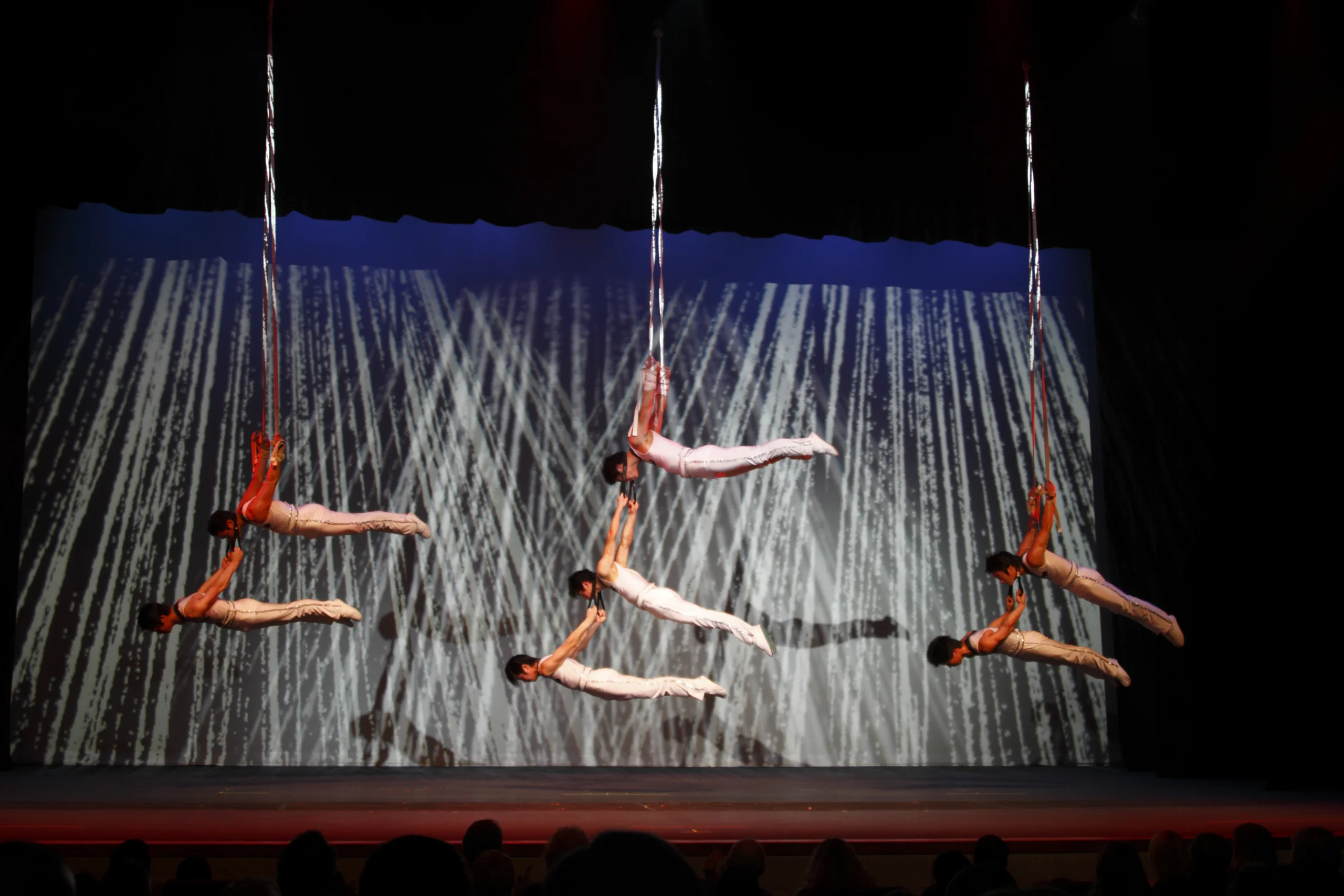 Acrobats perform body straightening in unison hanging from three ropes.