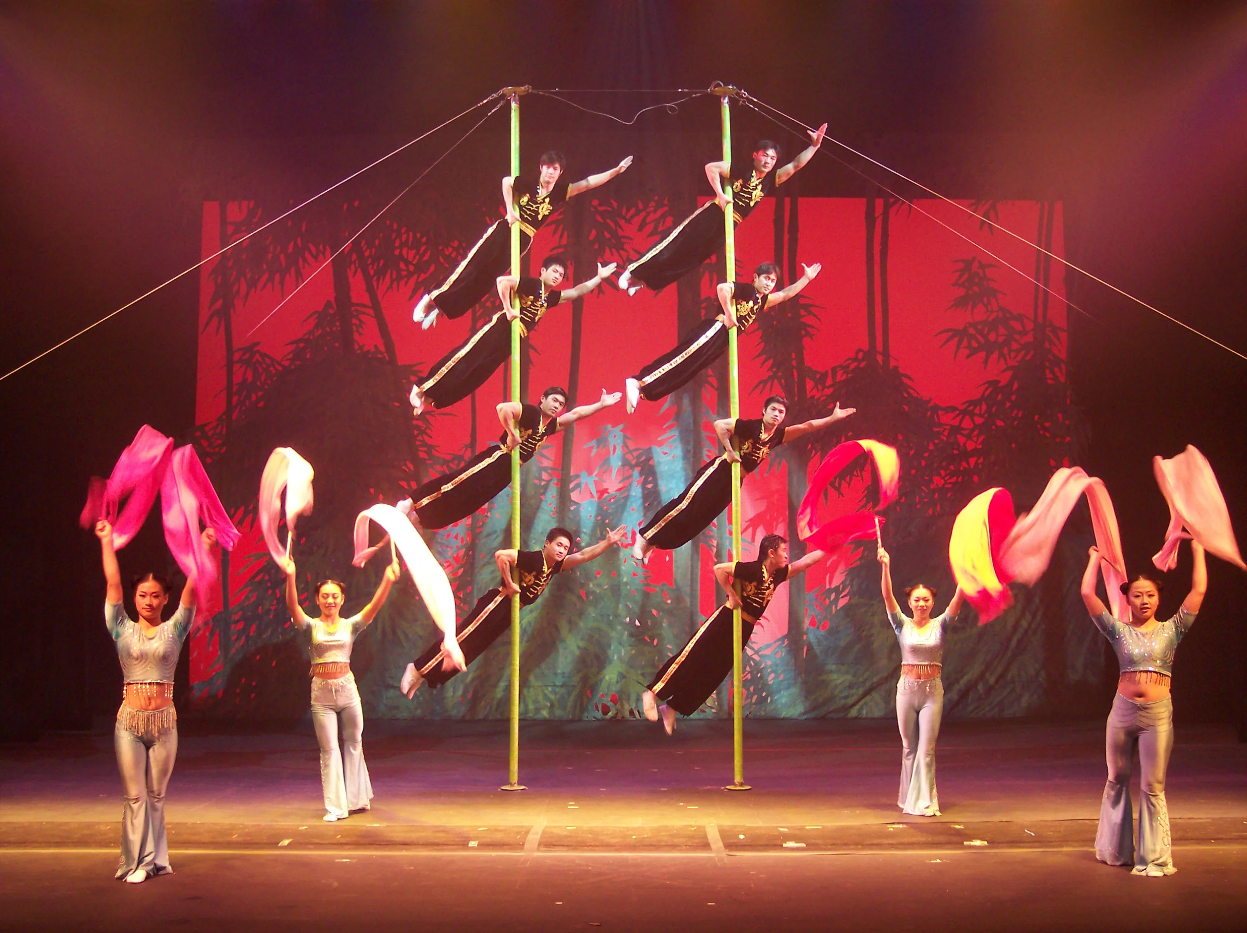 Acrobats in black and gold outfits hang horizontally from two vertical poles center stage, while four dancers in light blue costumes wave vibrant fabric ribbons below, set against a red and black forest-themed backdrop with dramatic lighting.