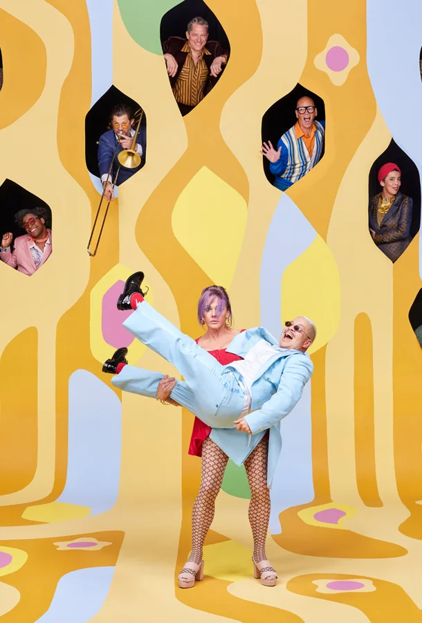 Members of Pink Martini poking their heads through different shaped holes on a colourful orange and yellow flat background and two performers in the middle, one holding the other cradle style.