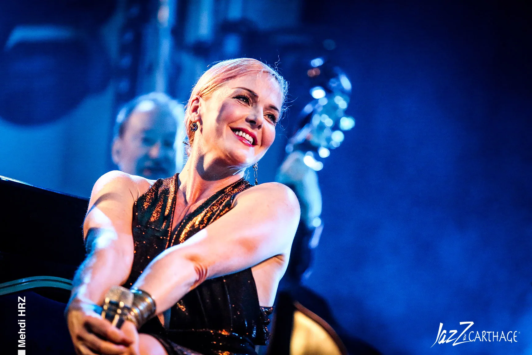 Blonde woman sitting on stage smiling resting a microphone by her knees.