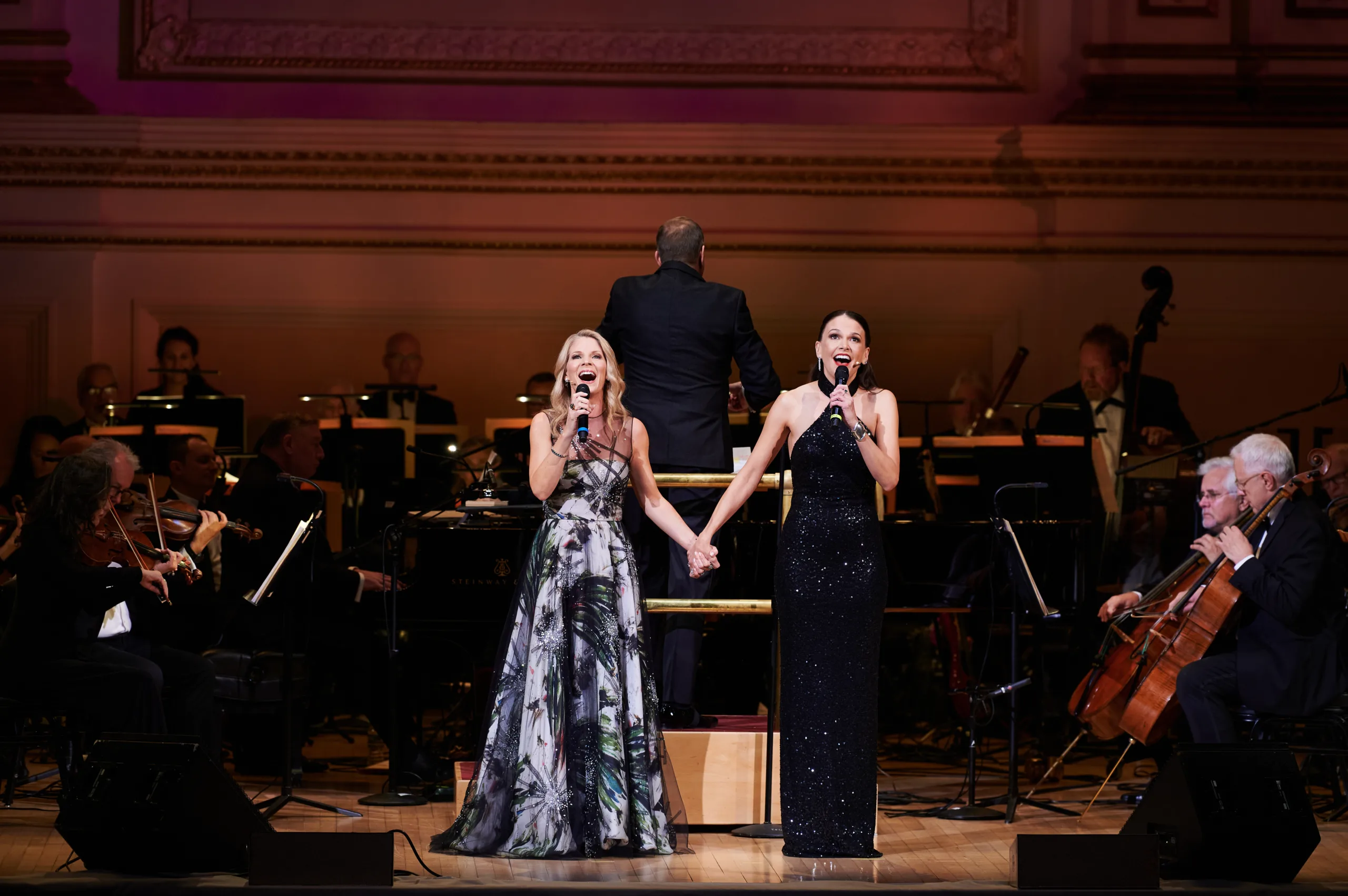 Sutton & Kelli holding hands singing on stage with orchestra behind them.
