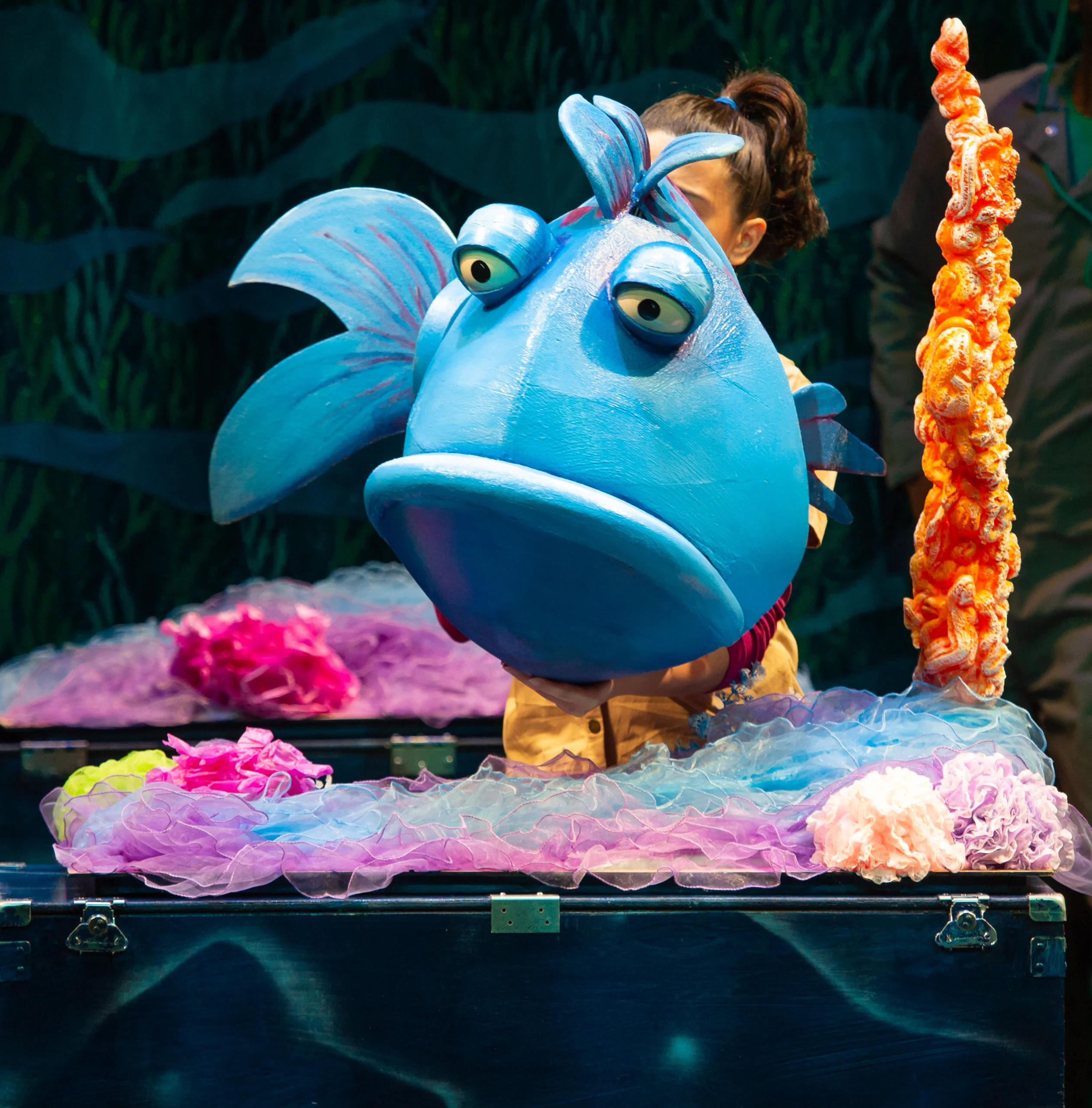 A person holding a large blue fish puppet in front of a colorful underwater-themed backdrop with coral and sea plant decorations