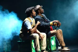 Actors on stage in a make believe car ,looking off into the distance
