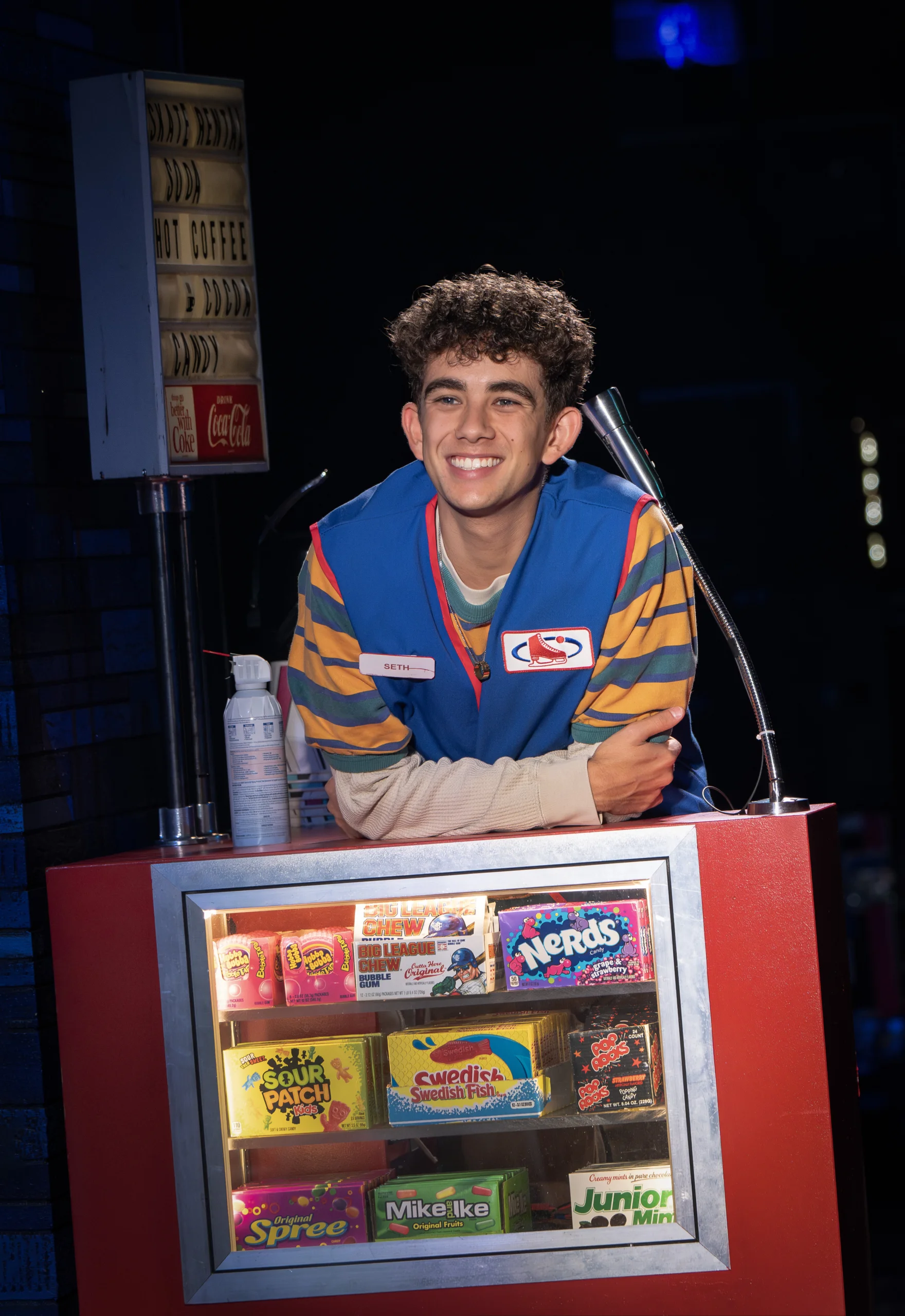 Teenage boy leaning on a concessions counter wearing a blue work vest and smiling.