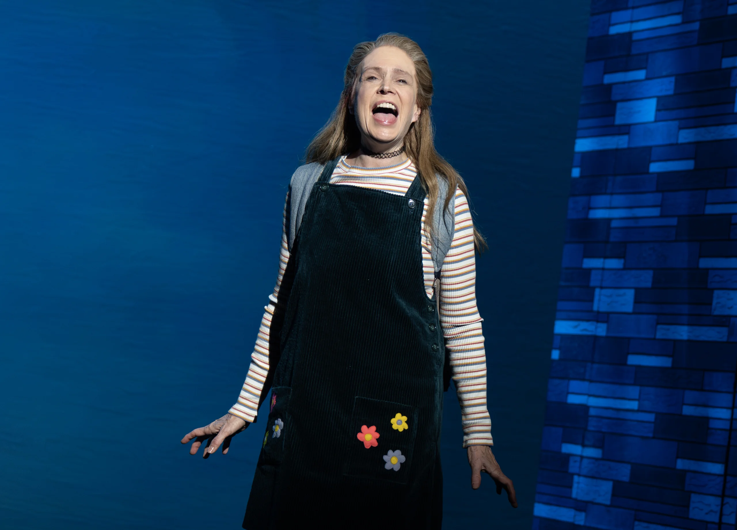Kimberly standing on stage singing in a pinafore dress and striped long-sleeved shirt. 