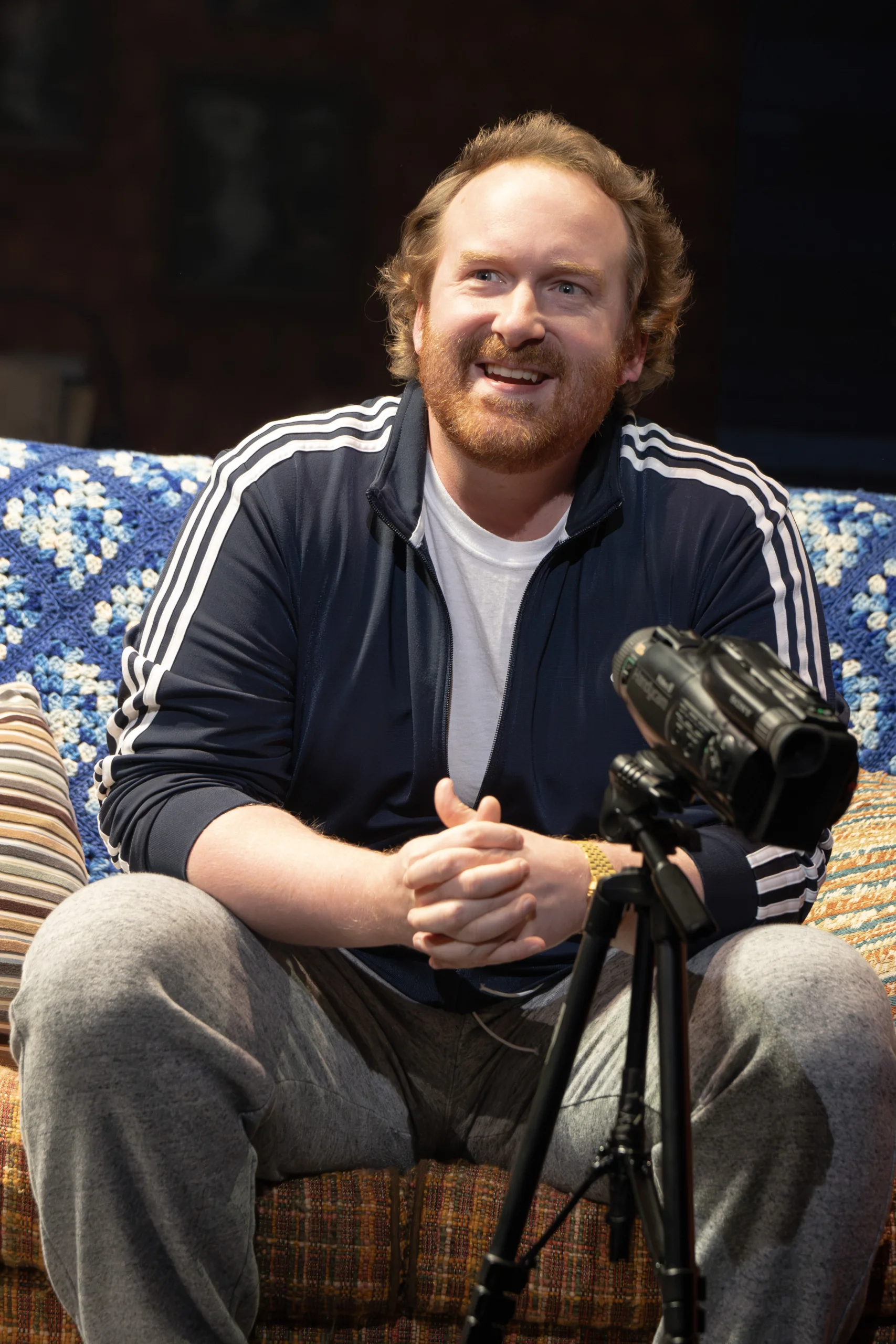 Buddy sitting on the couch wearing a sweatsuit in front of a video camera on a tripod.