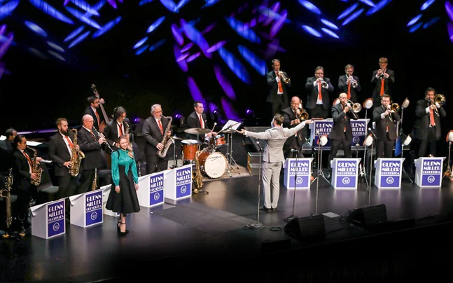 The Glenn Miller Orchestra performing on stage with musicians playing saxophones, trumpets, trombones, and drums. A conductor stands in the center with arms raised, and a female vocalist is positioned to the left. Colorful lighting effects illuminate the background, and music stands labeled 'Glenn Miller Orchestra' are visible.
