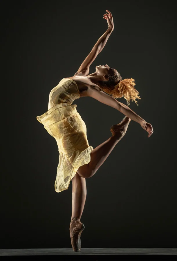 Ballet dancer in a flowing yellow dress performing an arabesque pose on pointe against a dark background.