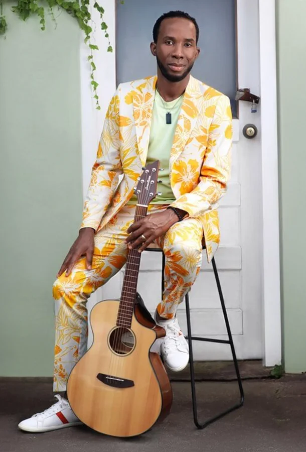 BélO in yellow suit poses on a stool with guitar.