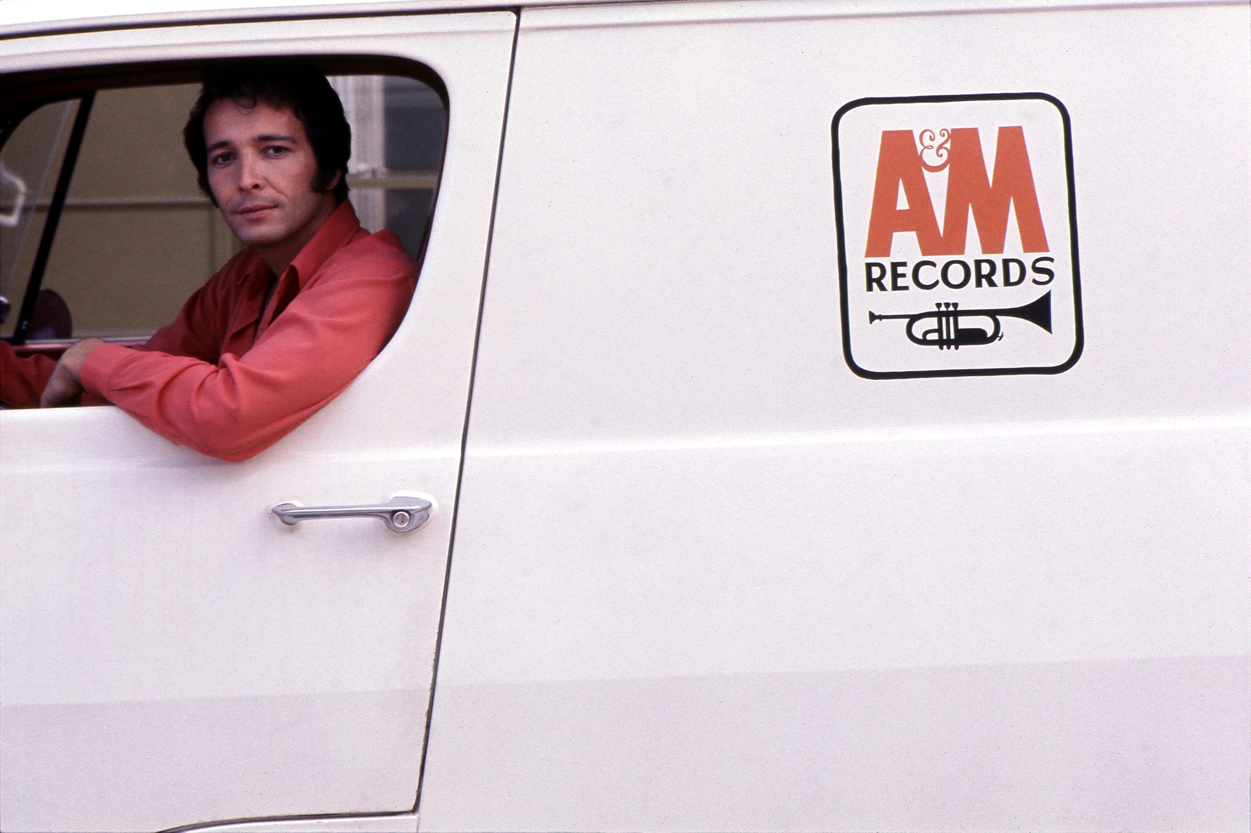 Herb Albert in a Am Records white van posing wearing red shirt.