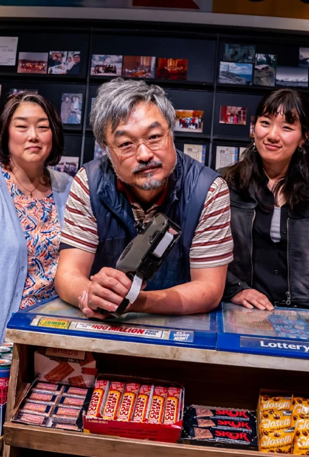 Mr. Kim leaning on the convenience counter holding a price gun with a woman over each shoulder and candies on display.